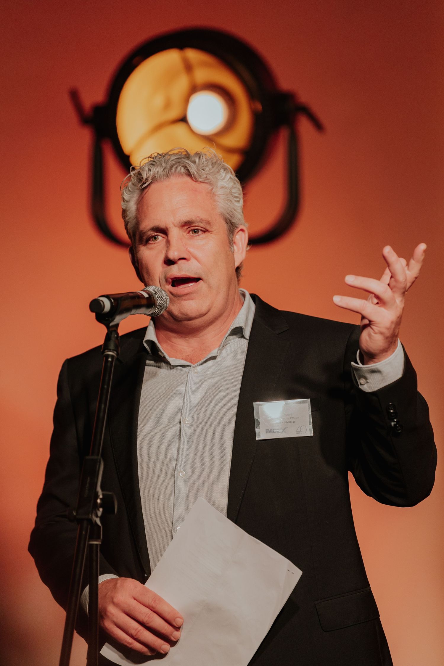 Middle-aged man in blazer speaking at a microphone on stage with vintage spotlight behind him.