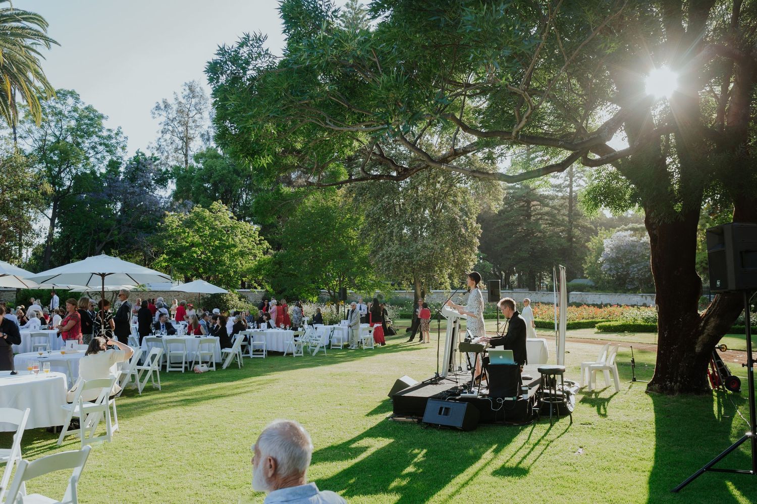 Outdoor garden wedding reception with white chairs and tables on green lawn, DJ setup under a large shady tree.