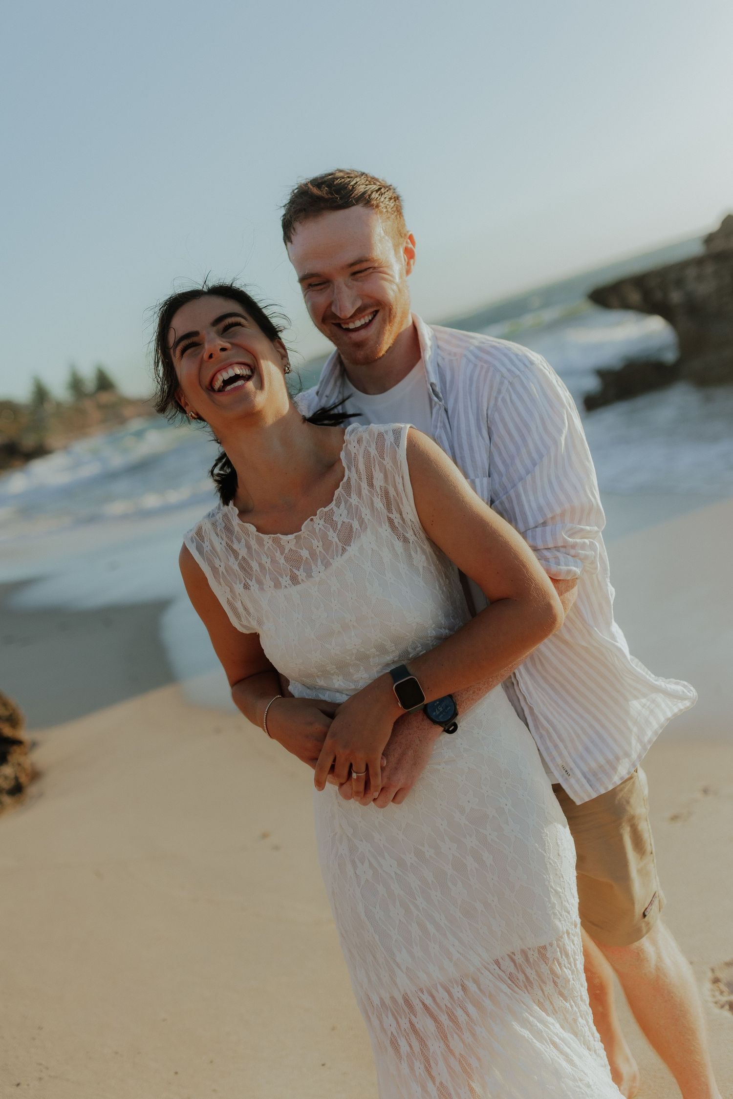 Joyful couple laughing together on a beach at sunset, woman in white lace dress embraced from behind by man.