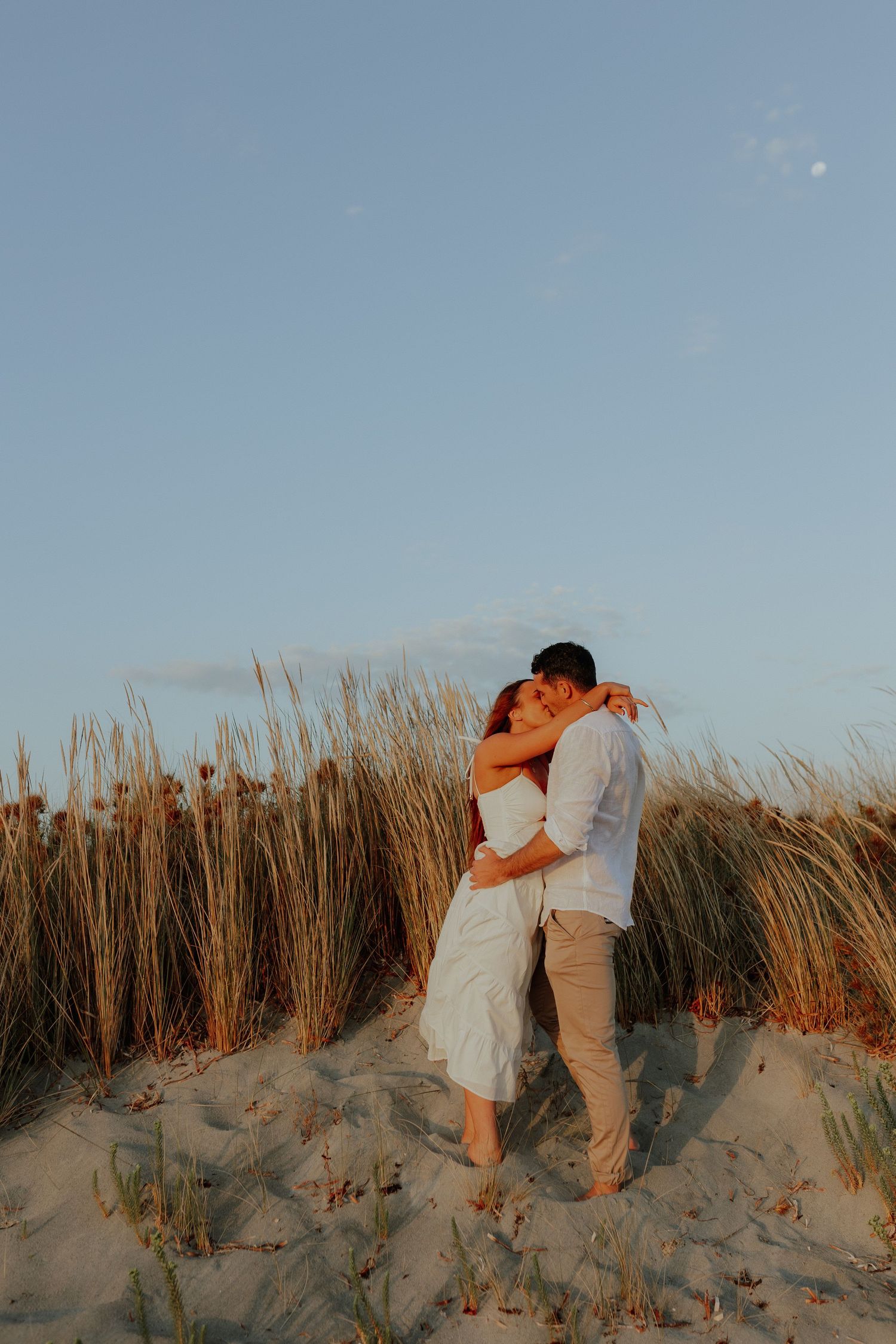 Couple kissing among golden beach grass dunes at sunset in white outfits.