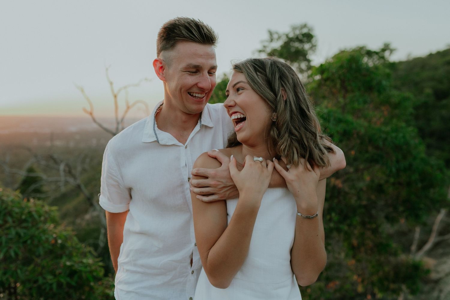 Happy couple embracing and laughing outdoors with lush green trees in the background at sunset.