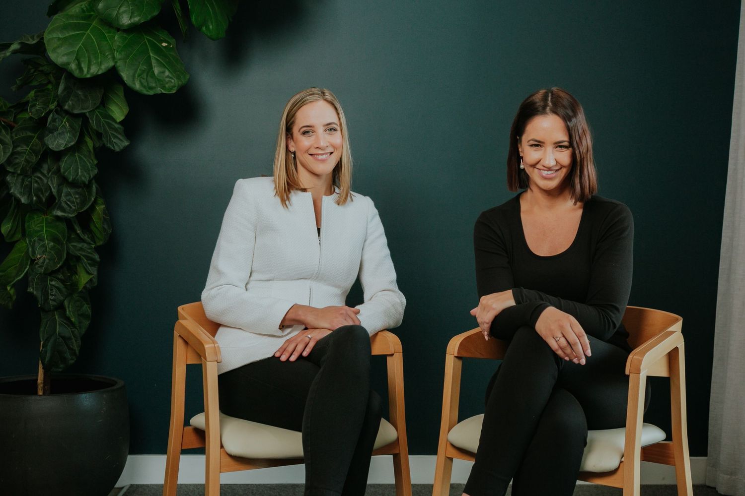 Two professional women smiling seated in wooden chairs against a dark teal wall.