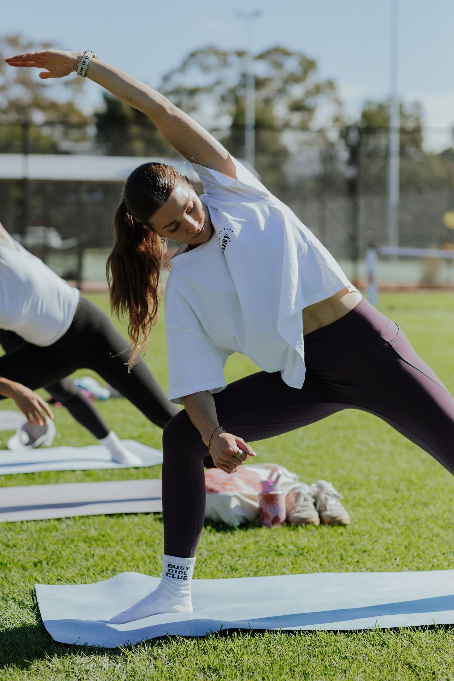 Woman in white top and leggings performing a yoga side stretch pose on a mat outdoors on green grass.