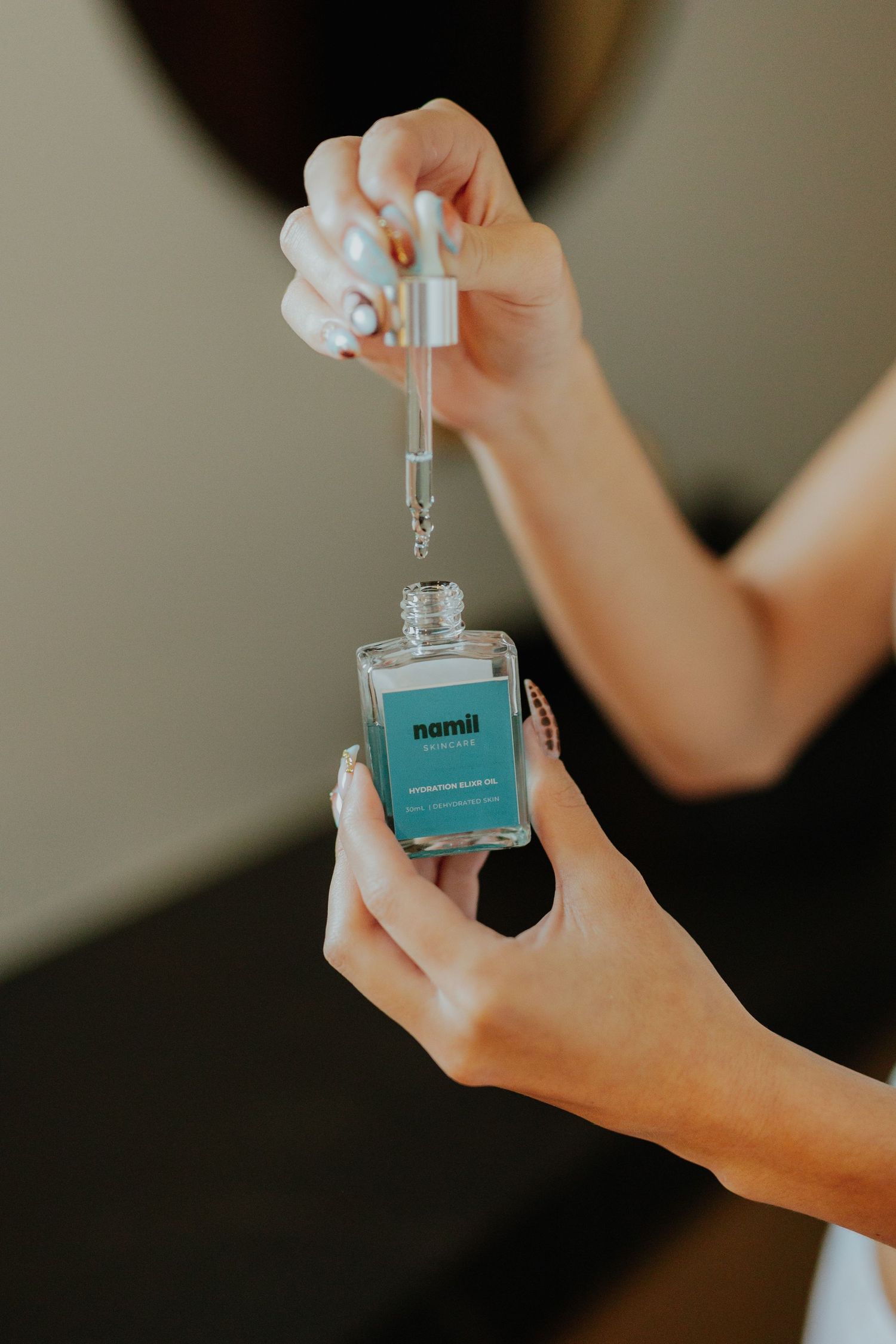 Hands holding a teal Nomii hydrating facial oil bottle with dropper against a neutral background.