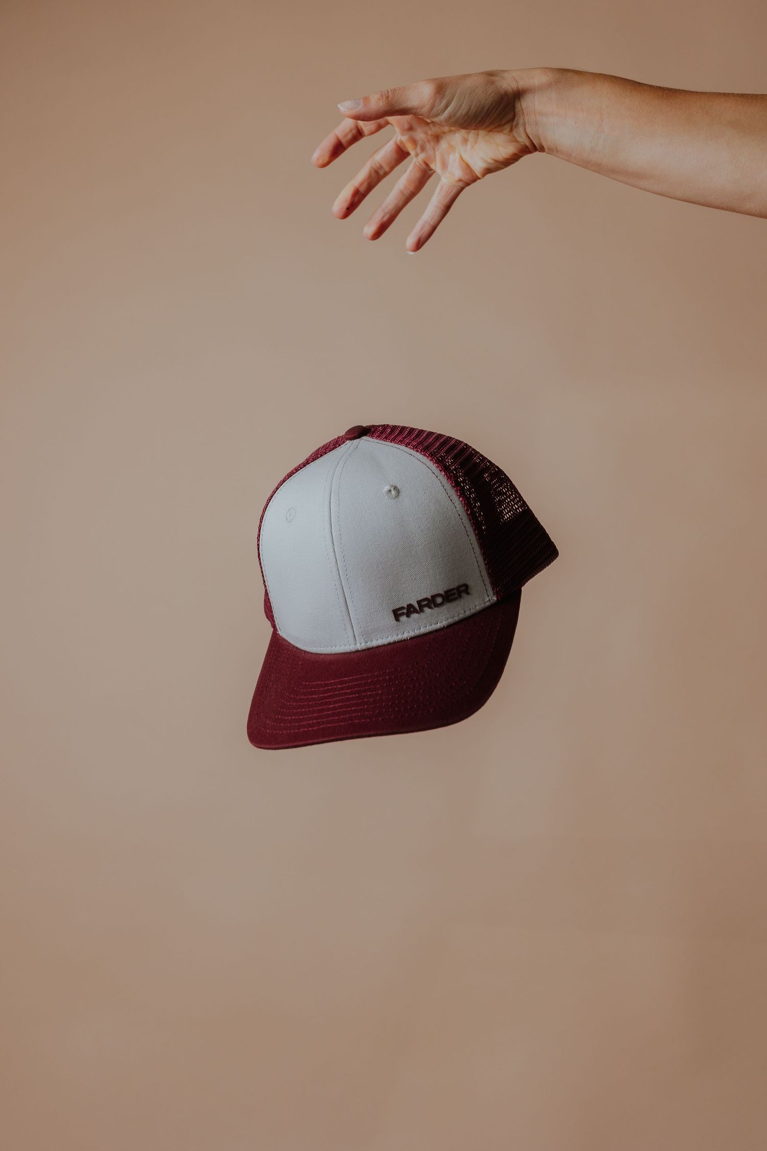 Maroon and white trucker cap with Farcer logo floating in mid-air against beige background with hand above.