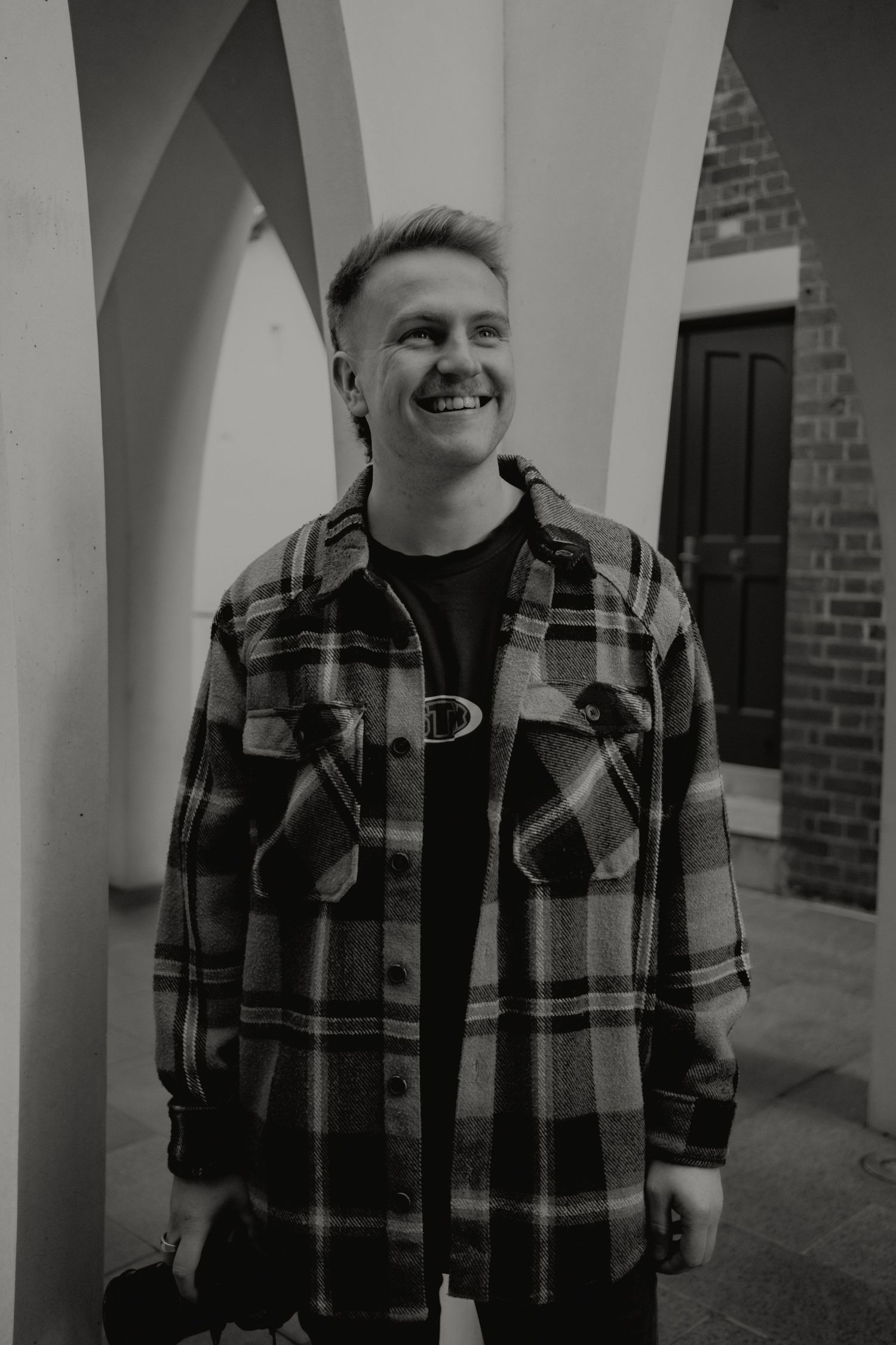Smiling man in plaid flannel shirt standing between white arched columns in black and white.