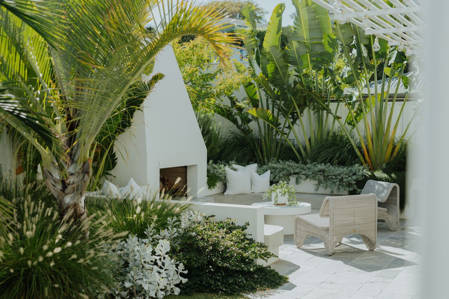 Lush tropical garden with white outdoor furniture and a fireplace surrounded by palm trees.