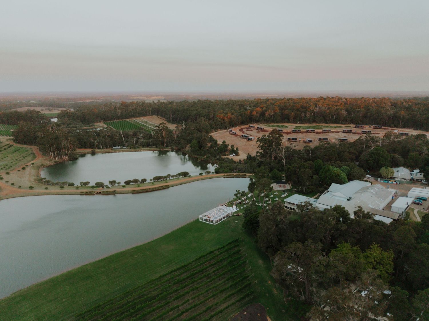 Aerial drone view of wedding venue surrounded by lake, green fields, and autumn trees at dusk.