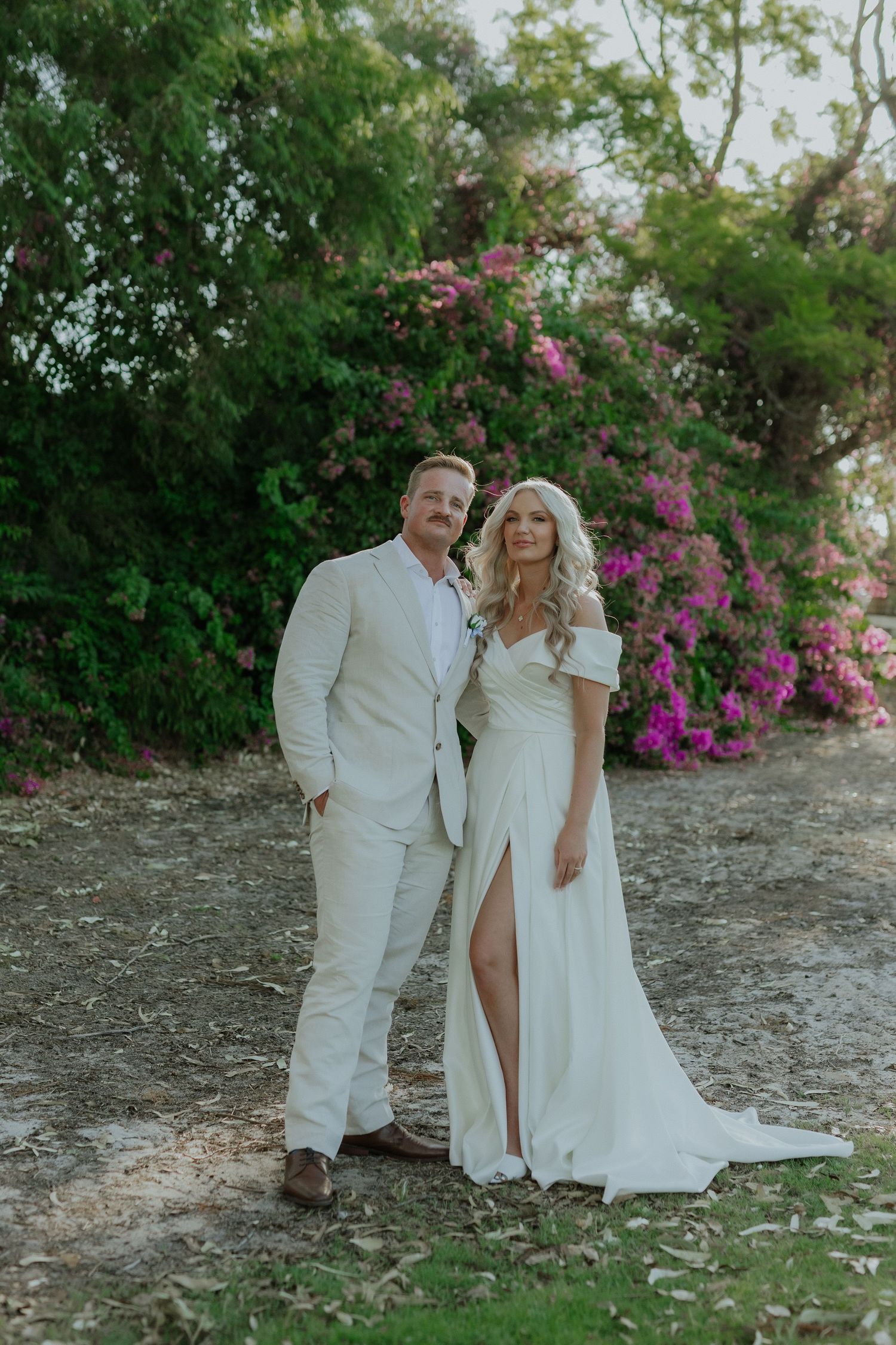 Bride in white slit gown and groom in cream suit posing before colorful garden blooms.