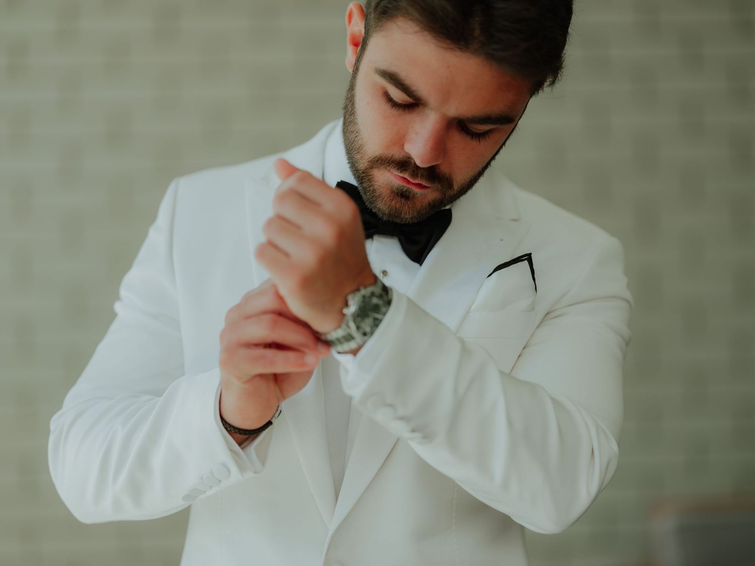 Groom in white tuxedo jacket and black bow tie adjusting his cufflinks while looking down at his wrist.