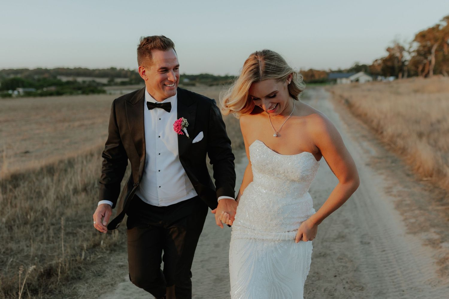 Smiling bride and groom walk hand-in-hand along a dusty country road in golden hour light.