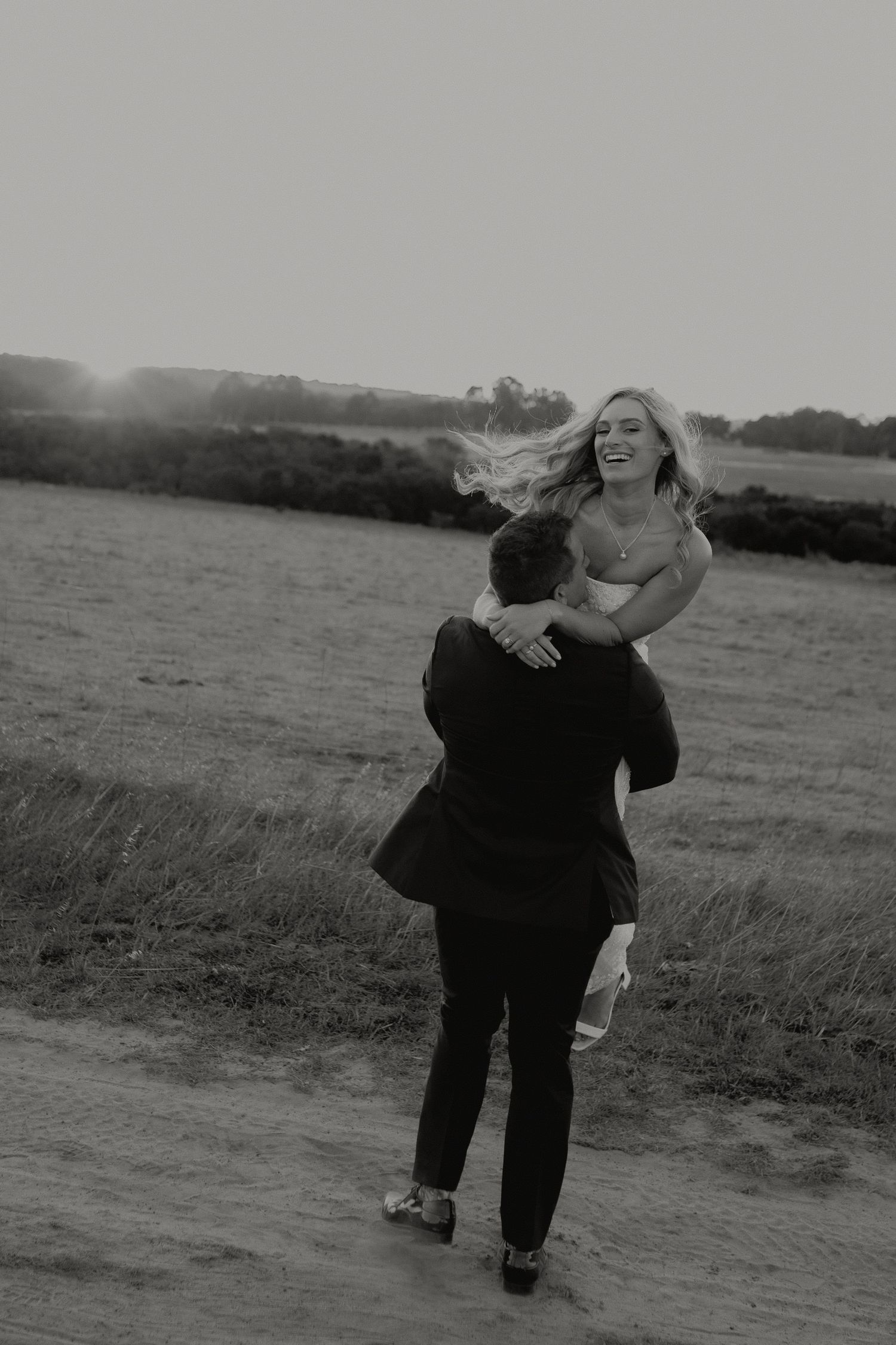 Black and white photo of groom lifting joyful bride in field at sunset during wedding portraits.