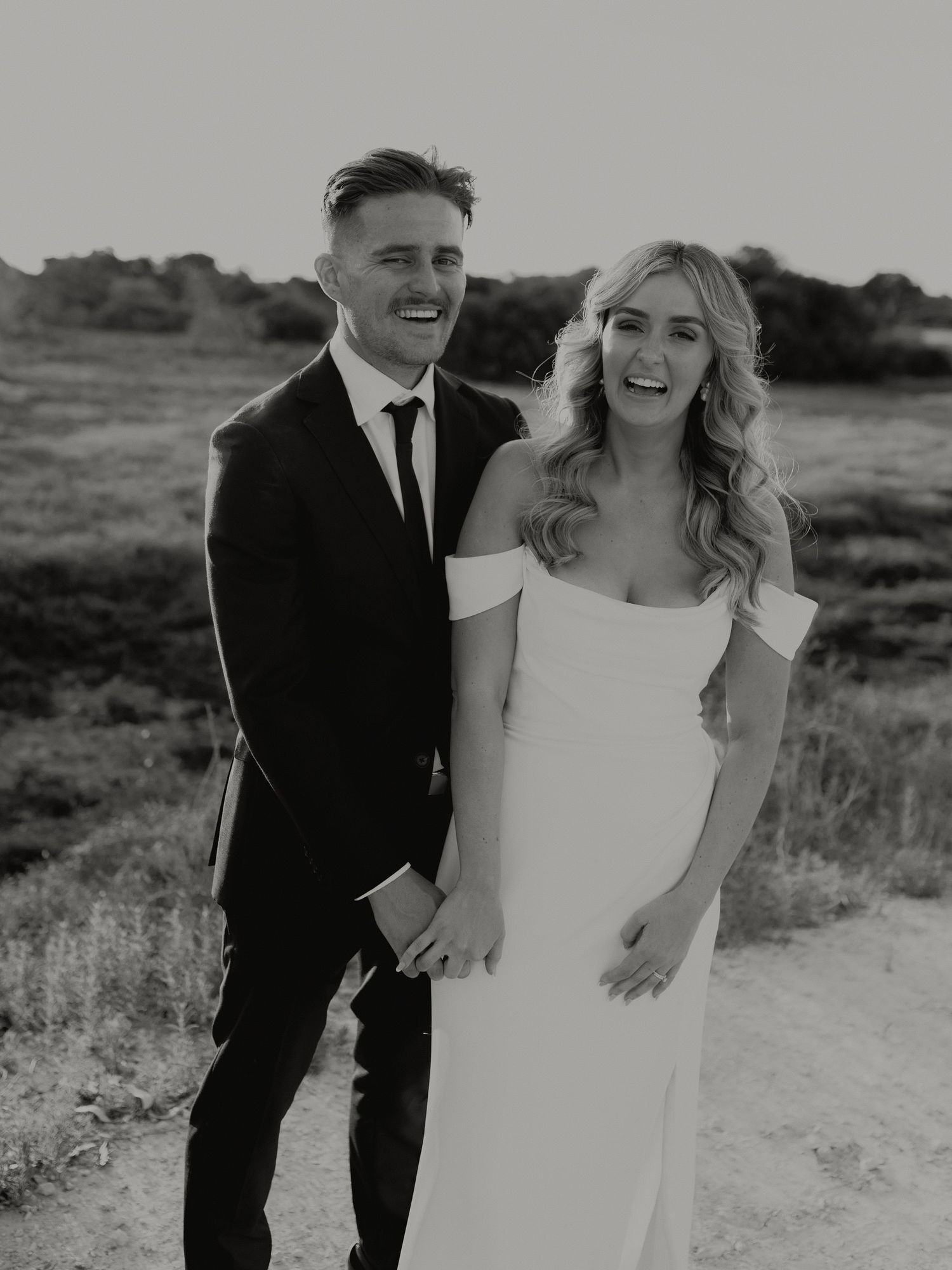 Black and white portrait of smiling bride and groom holding hands on a rural dirt road.