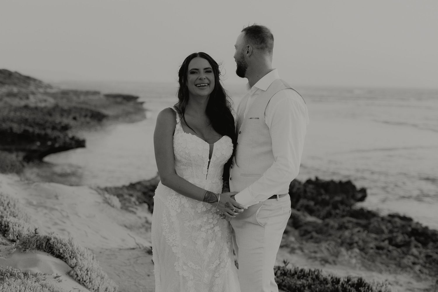 Black and white photo of bride and groom holding hands on rocky coastal cliffs at sunset.