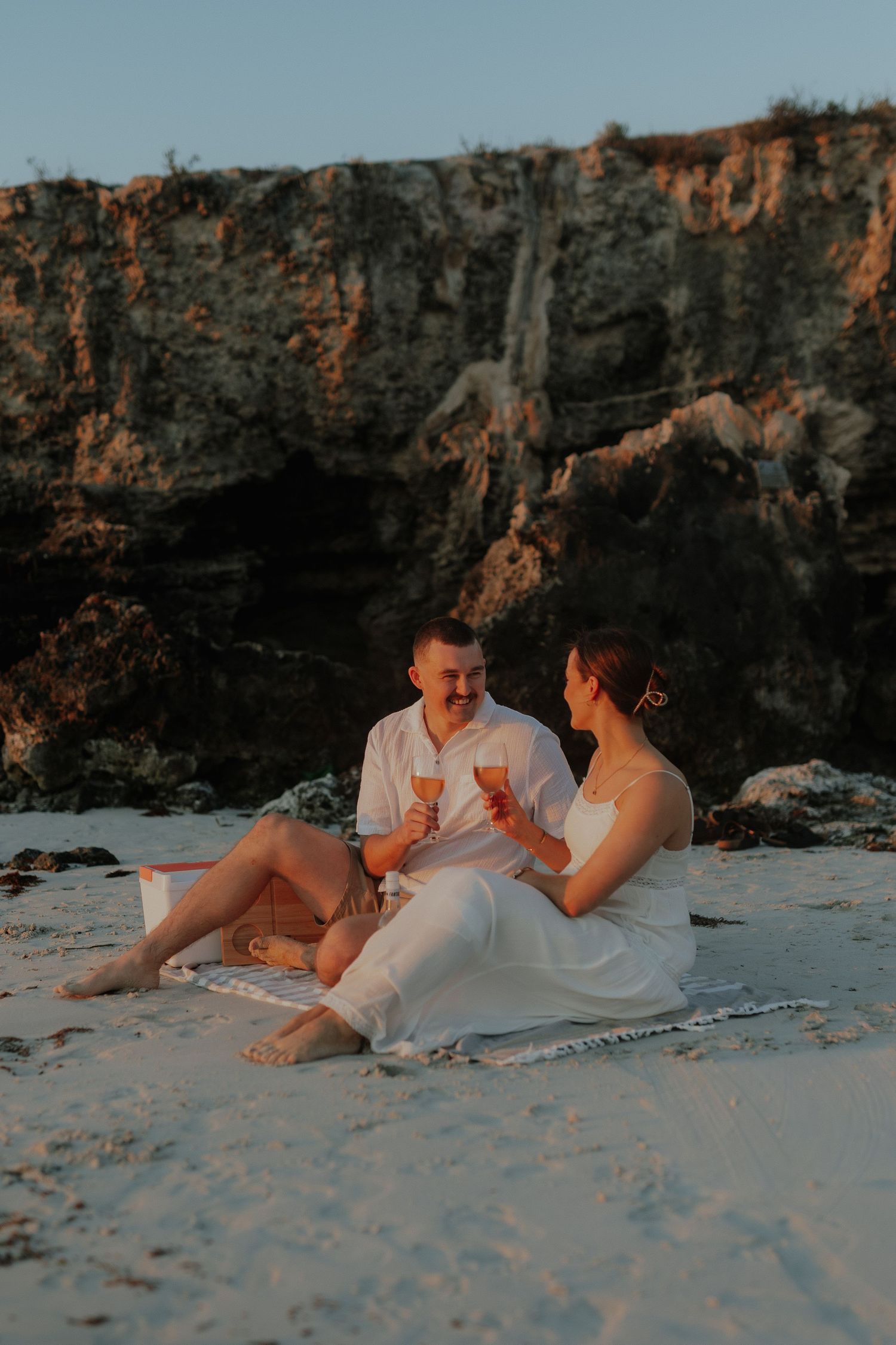 Happy couple sitting on white sand beach with wine glasses near rocky cliffs at sunset.