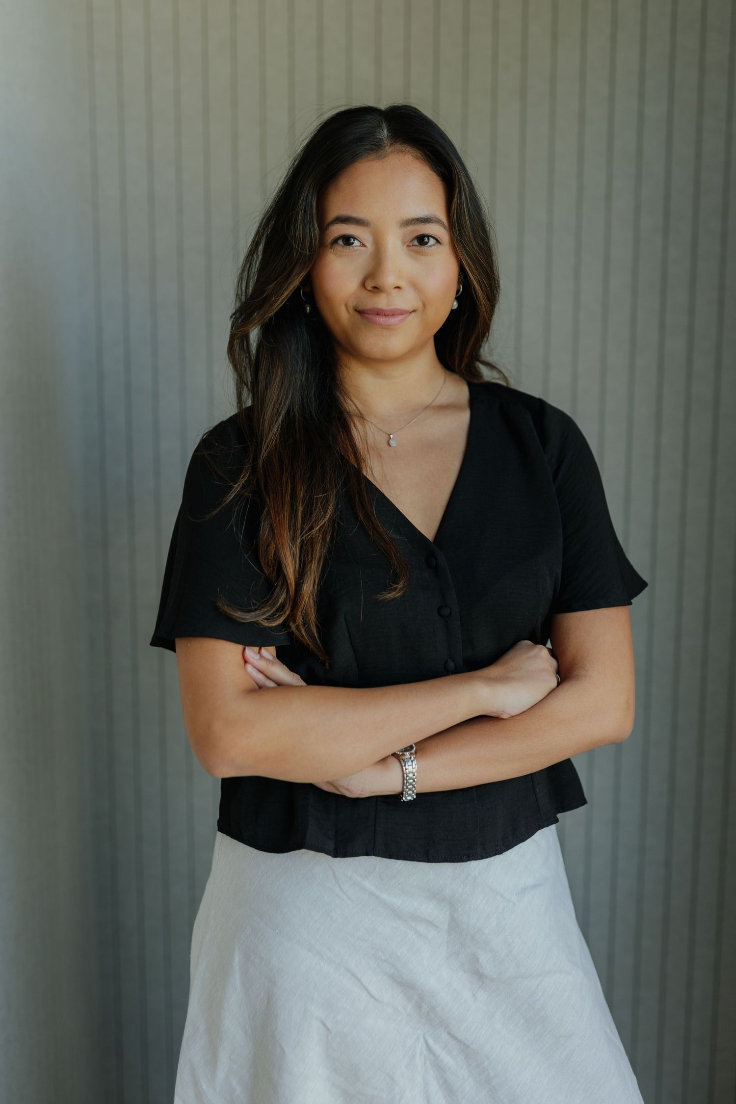 Young woman in black top and white skirt standing with arms crossed against a grey background.