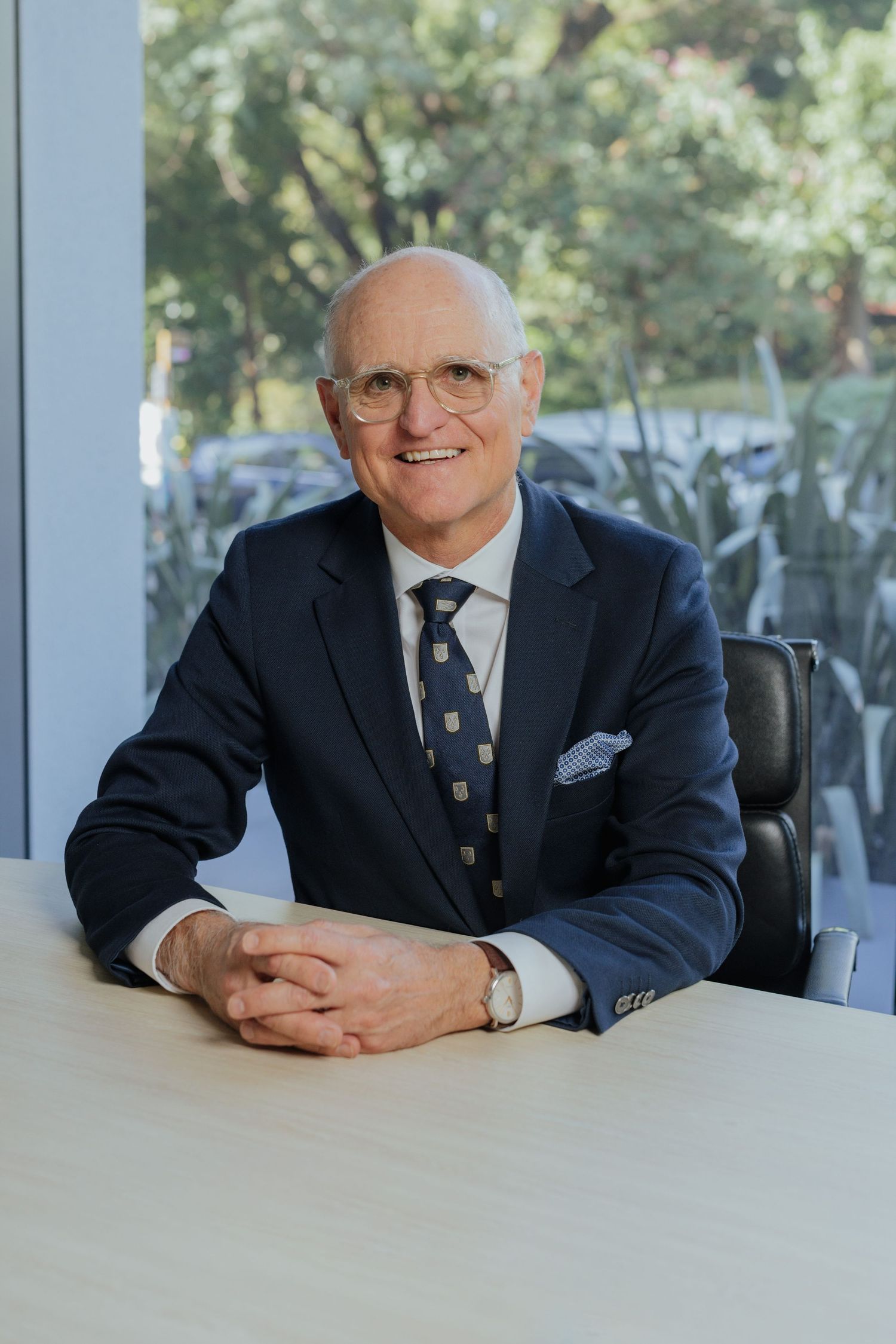 Senior male executive in navy suit smiles while seated at a desk in a modern office.