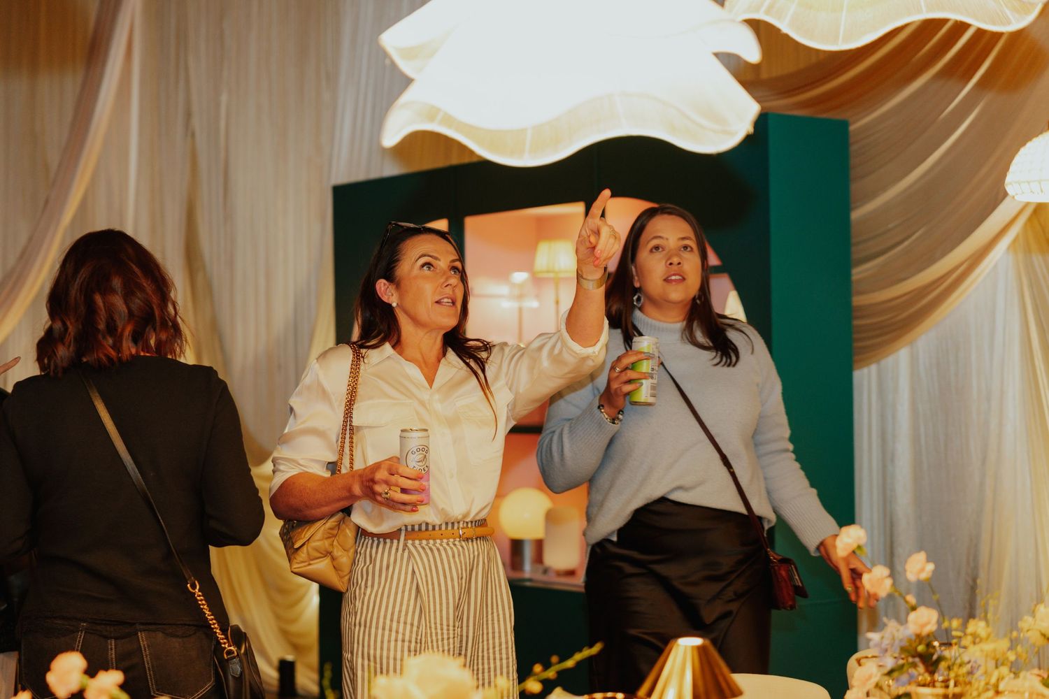 Two women smile and gesture at an elegant indoor event with white draping and floral decorations.