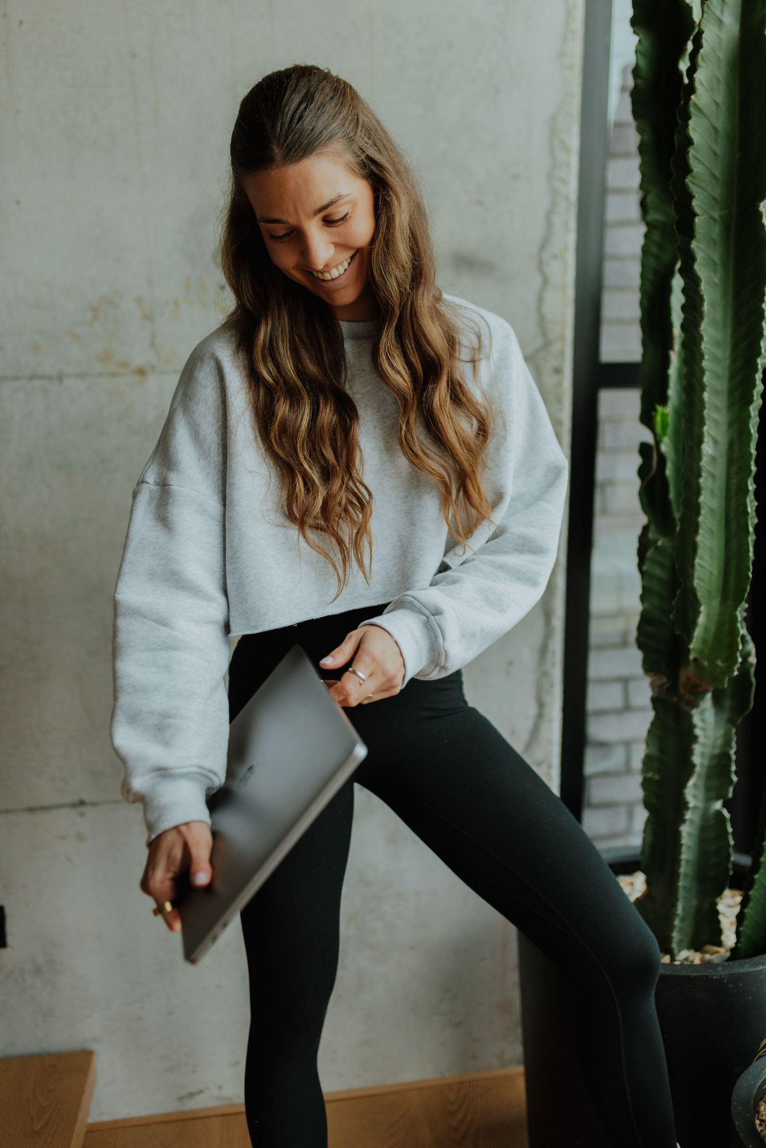 Laughing woman in grey crop sweatshirt and black leggings looking down at laptop near cactus.