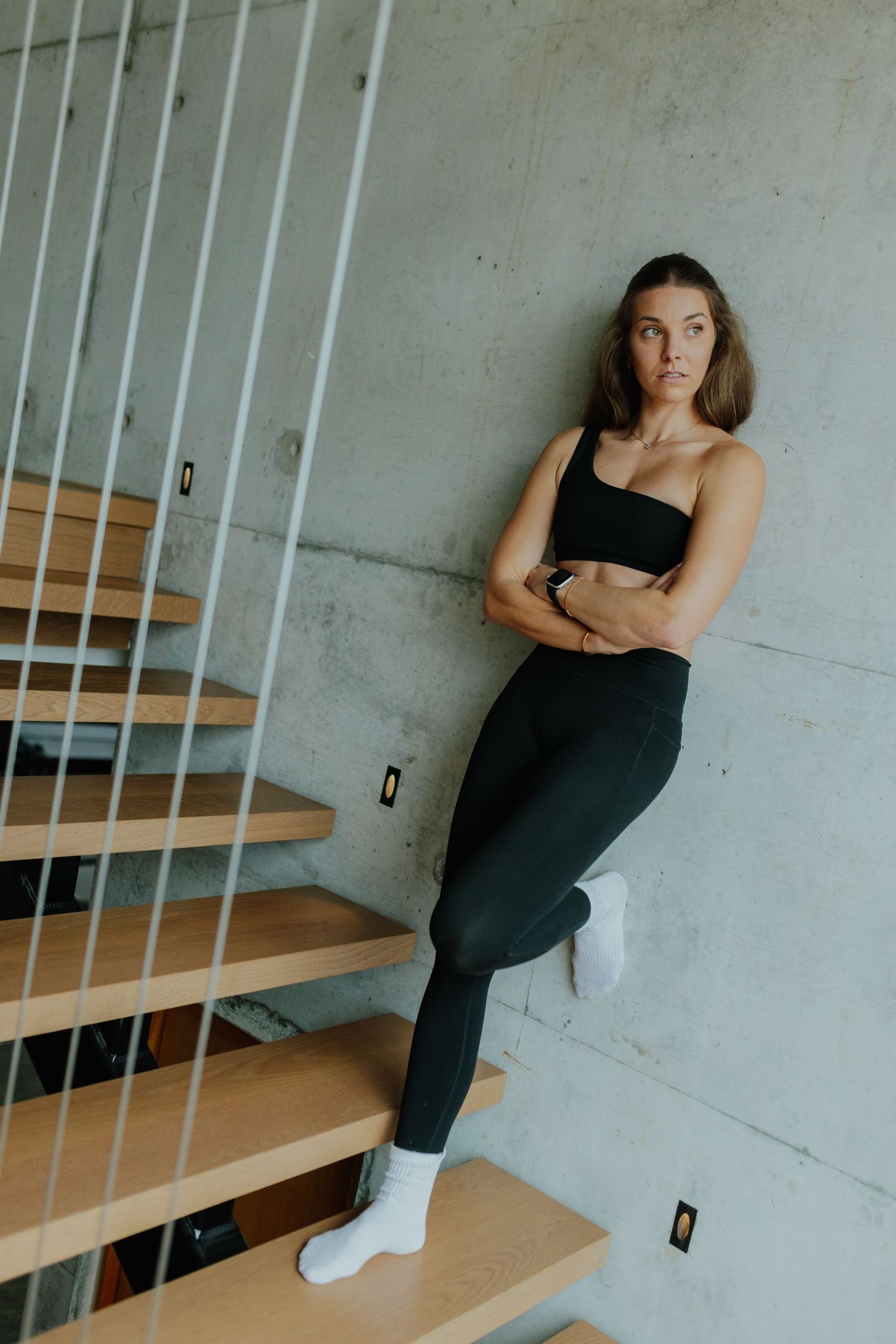 Athletic woman in black sports bra and leggings leaning against concrete wall by wooden stairs.
