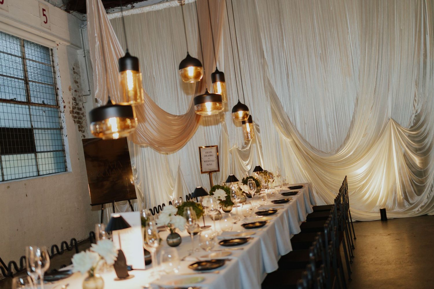 Elegant wedding reception table with dark pendant lights, black napkins, and white draping along the wall.