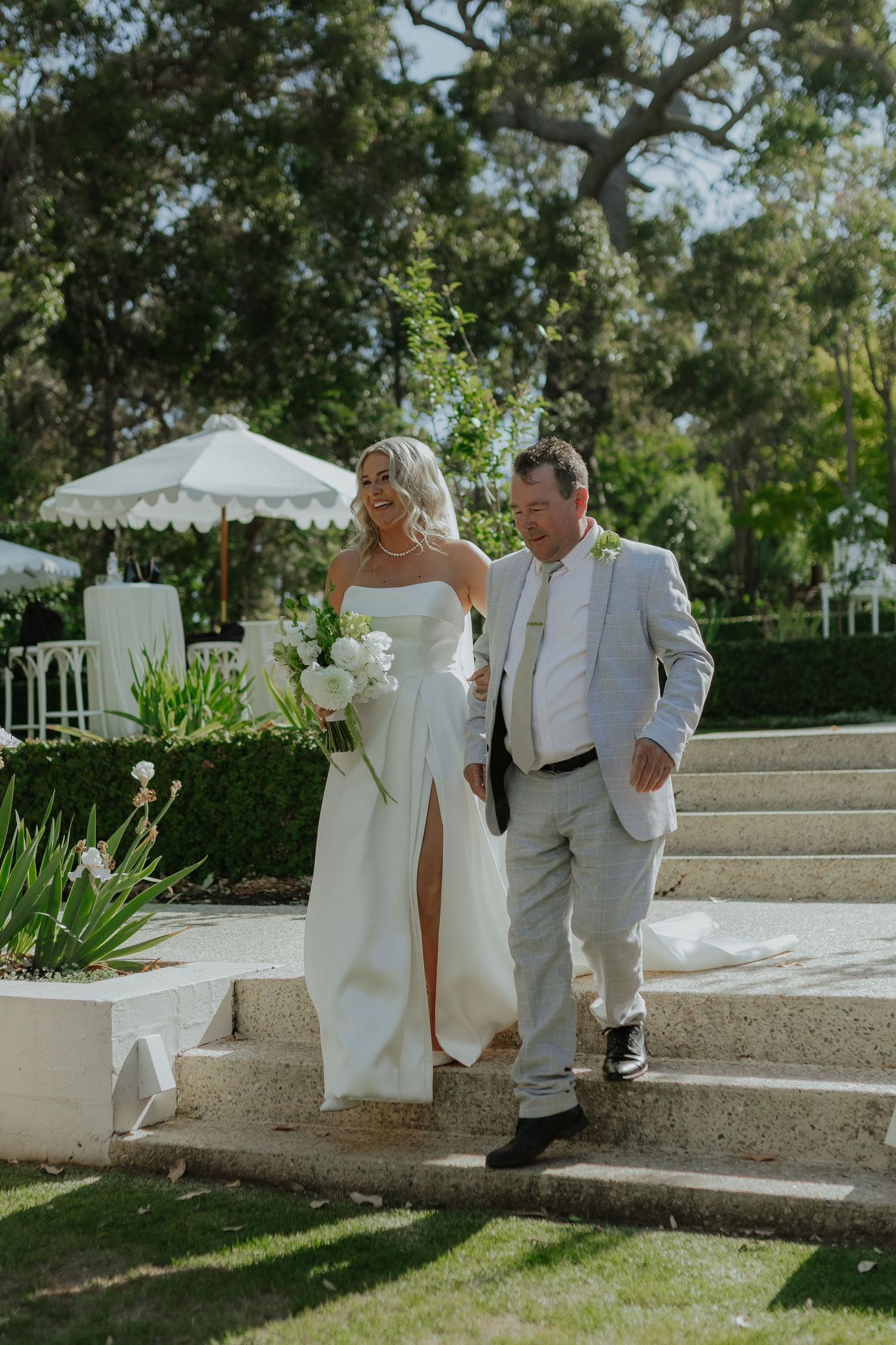 Bride in white strapless gown walks down stone steps with older man in gray suit at outdoor wedding.