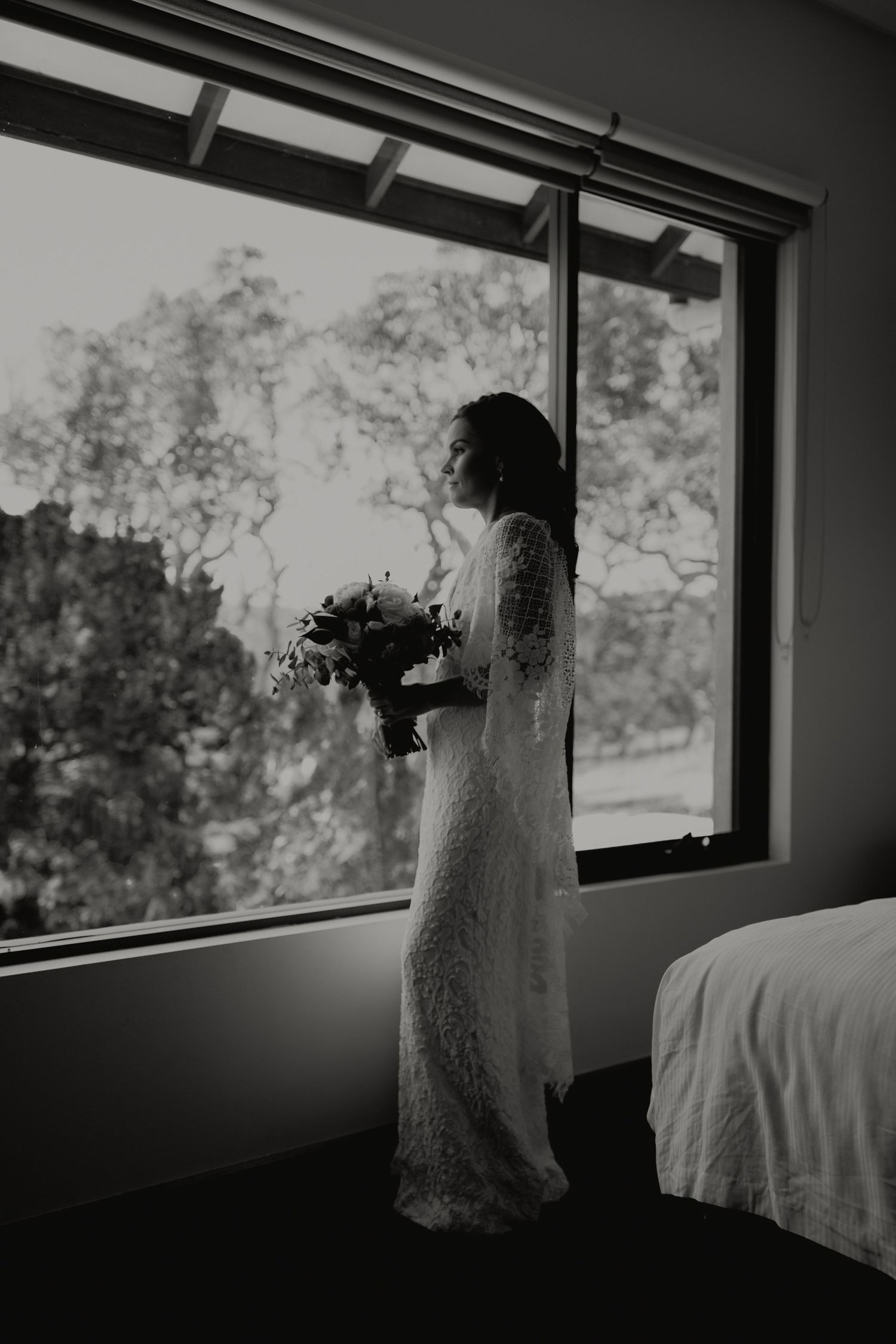 Bride in lace gown holds bouquet while gazing out large window in black and white portrait.