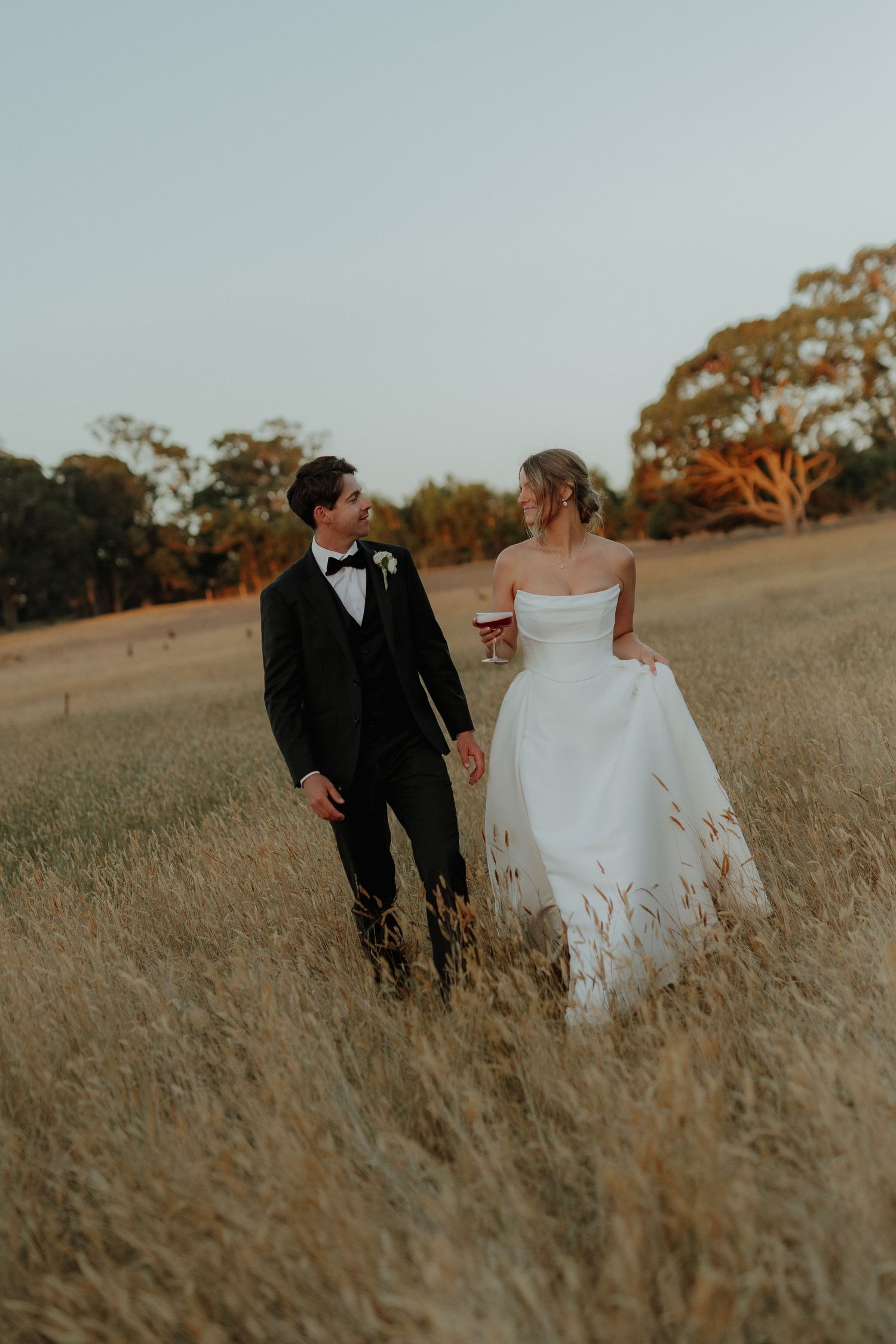 Bride and groom walking through golden wheat field at sunset, groom in black tuxedo, bride in strapless gown.