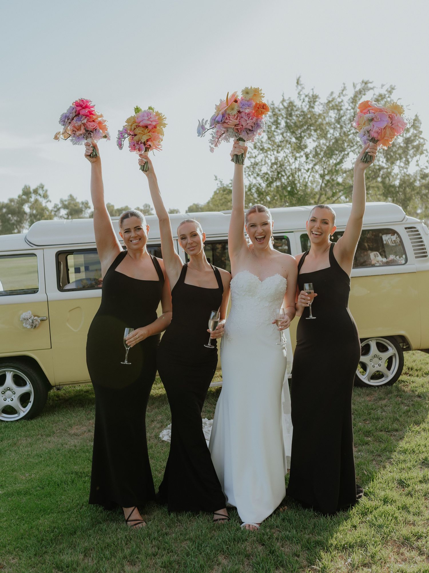 Bride with three bridesmaids in black gowns raising colorful bouquets beside vintage VW campervan.