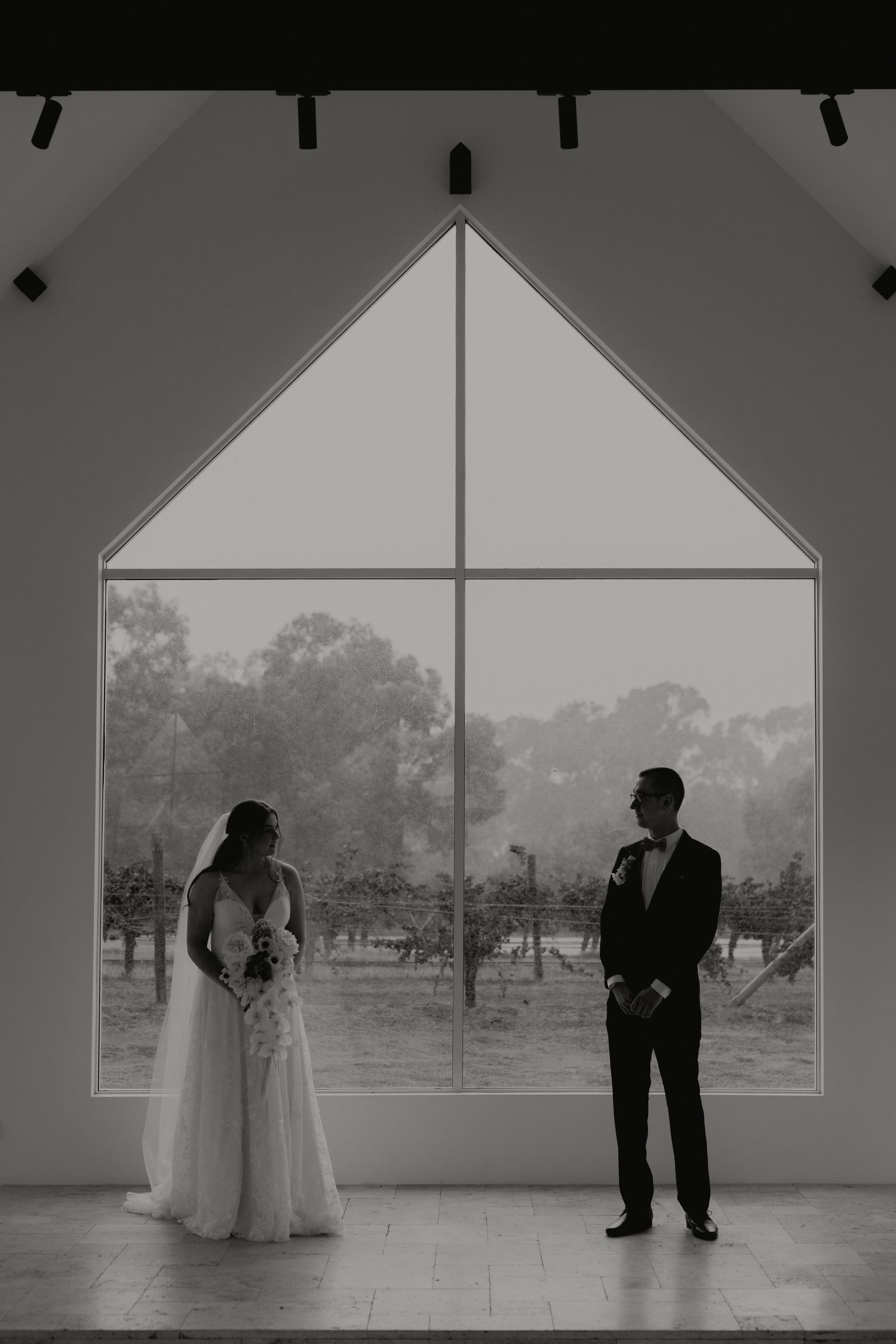 Bride and groom standing by large chapel window in black and white.