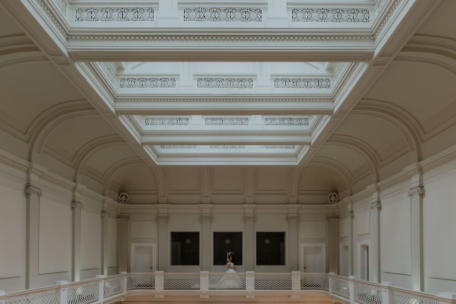 Elegant neoclassical ballroom with ornate white coffered ceiling, arched alcoves, and a lone figure in the center.