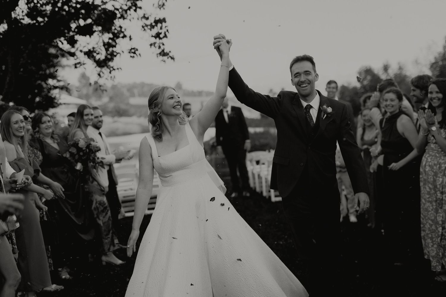 Black and white photo of smiling bride and groom walking through confetti shower at outdoor ceremony.