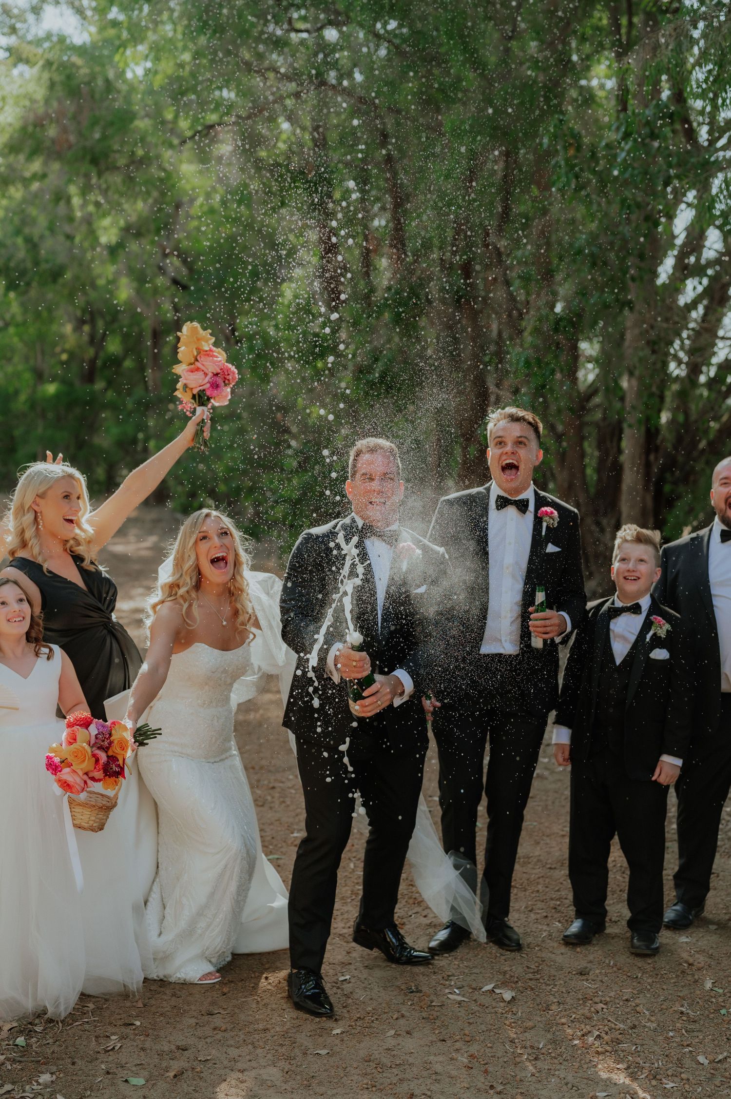 Wedding party celebrating with champagne spray outdoors, bride and groom in formal attire.