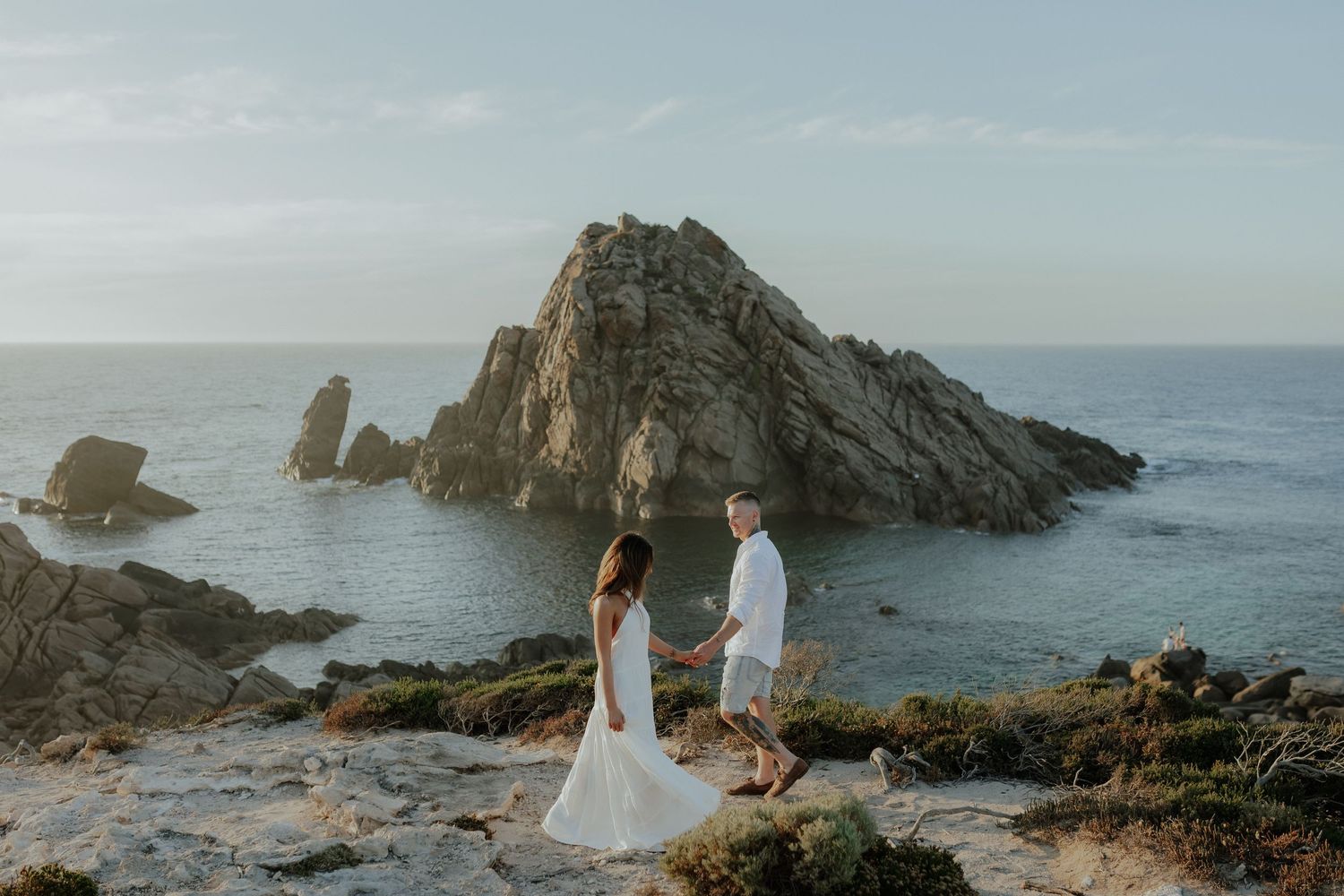 Couple holding hands on rocky coastal cliffs with a dramatic sea stack and ocean backdrop.