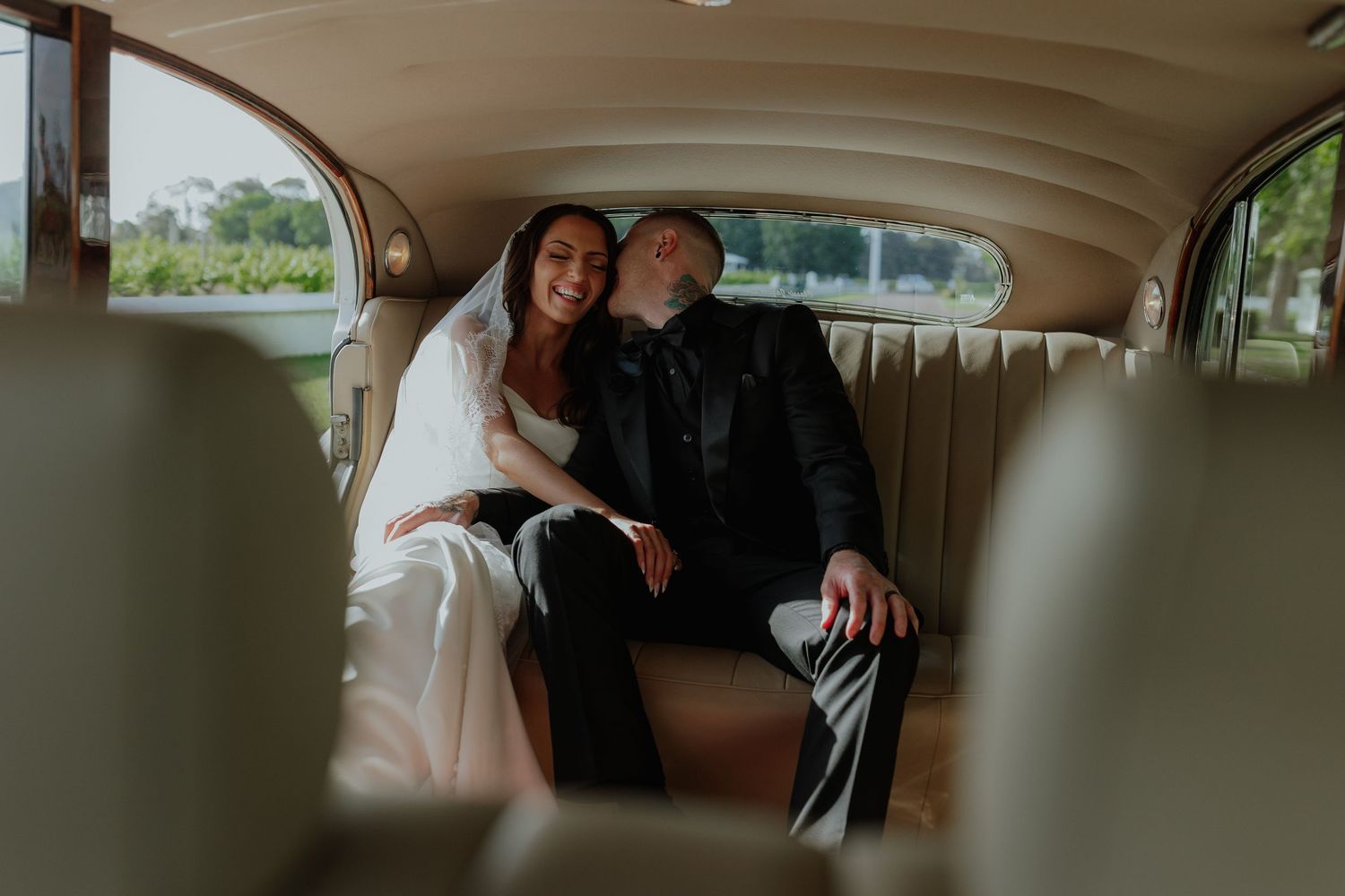 Bride and groom sitting together in vintage classic car backseat smiling.