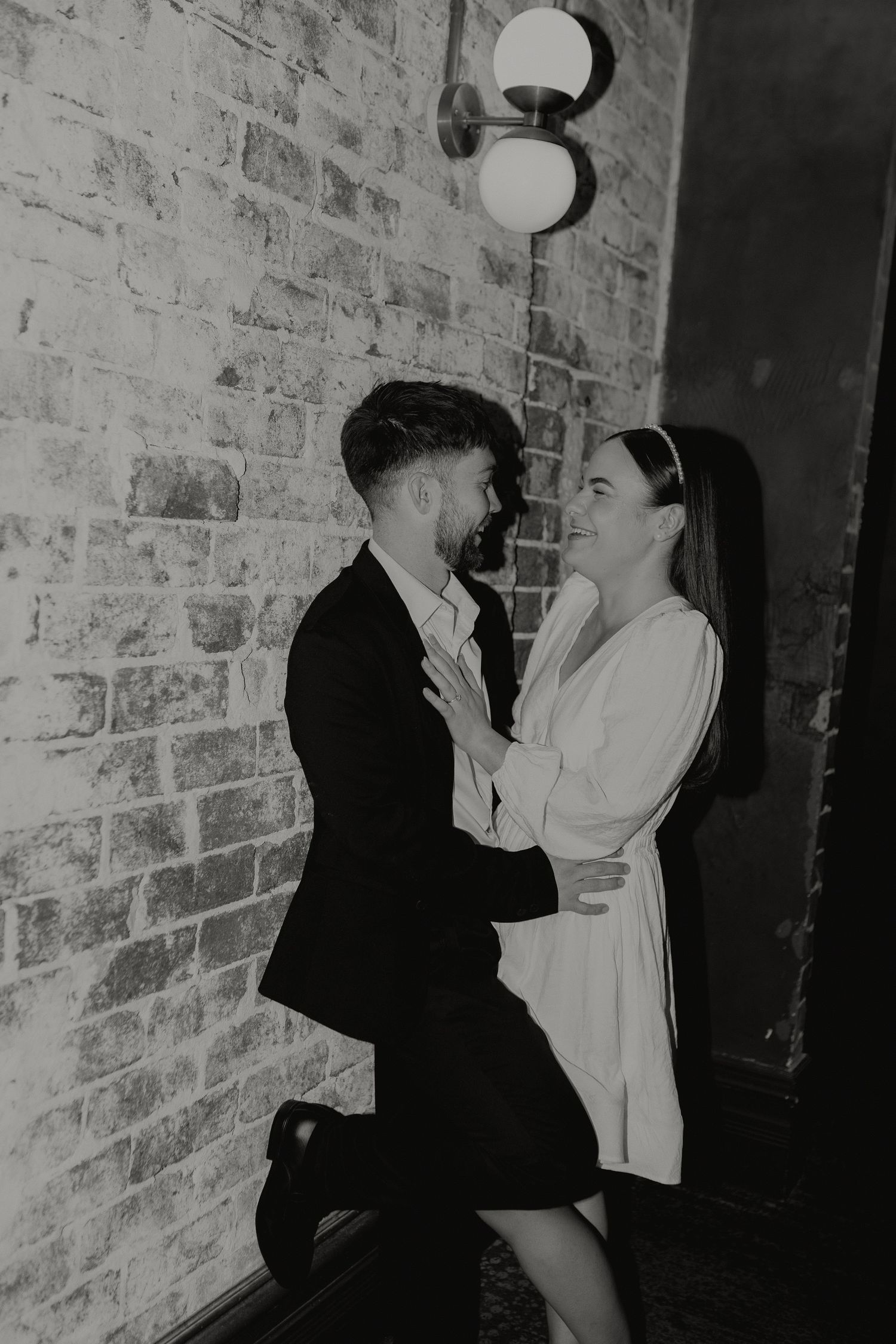 Black and white photo of a couple sharing a romantic moment against a brick wall indoors.