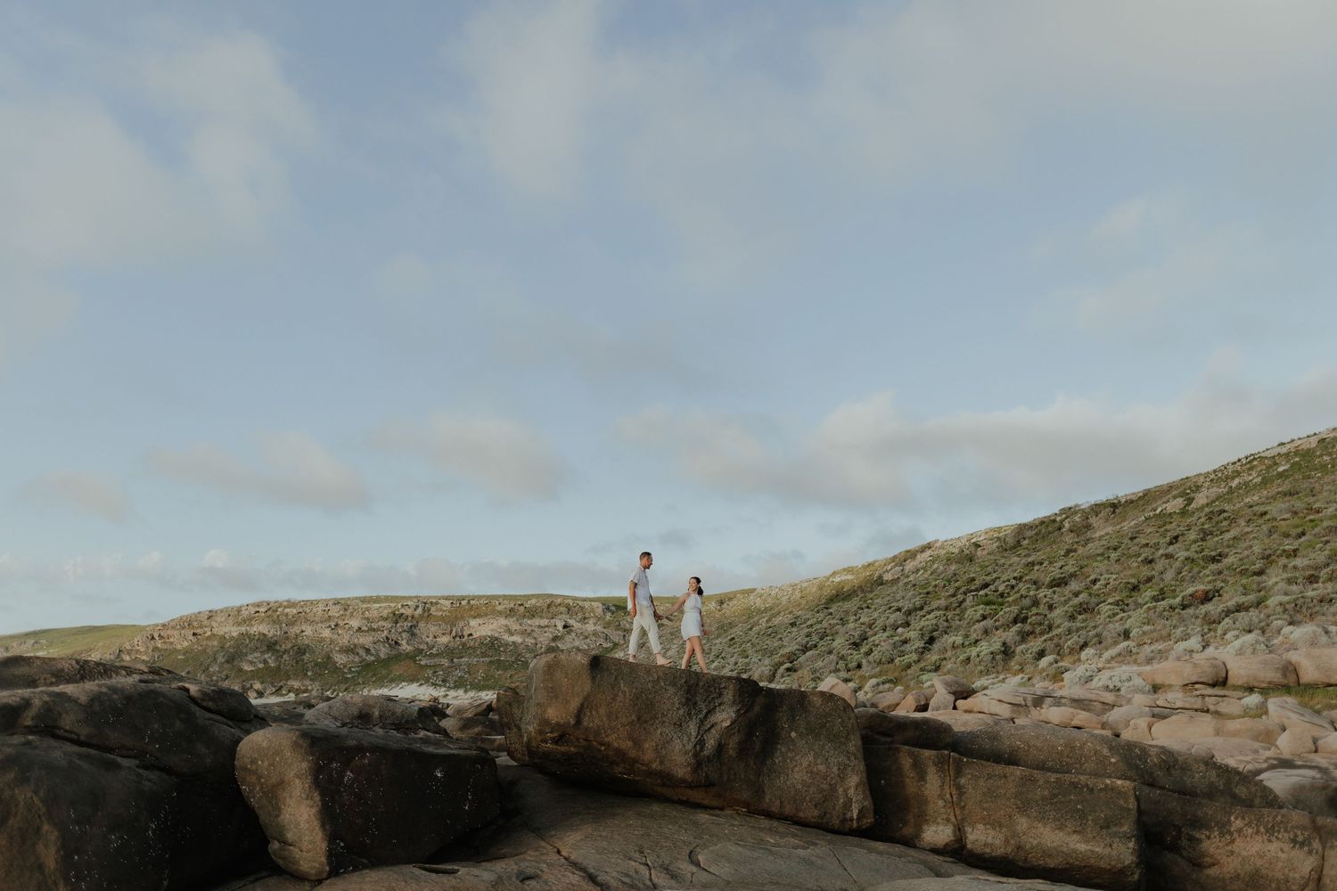 Couple standing on coastal rocks with green hills and cloudy sky backdrop.
