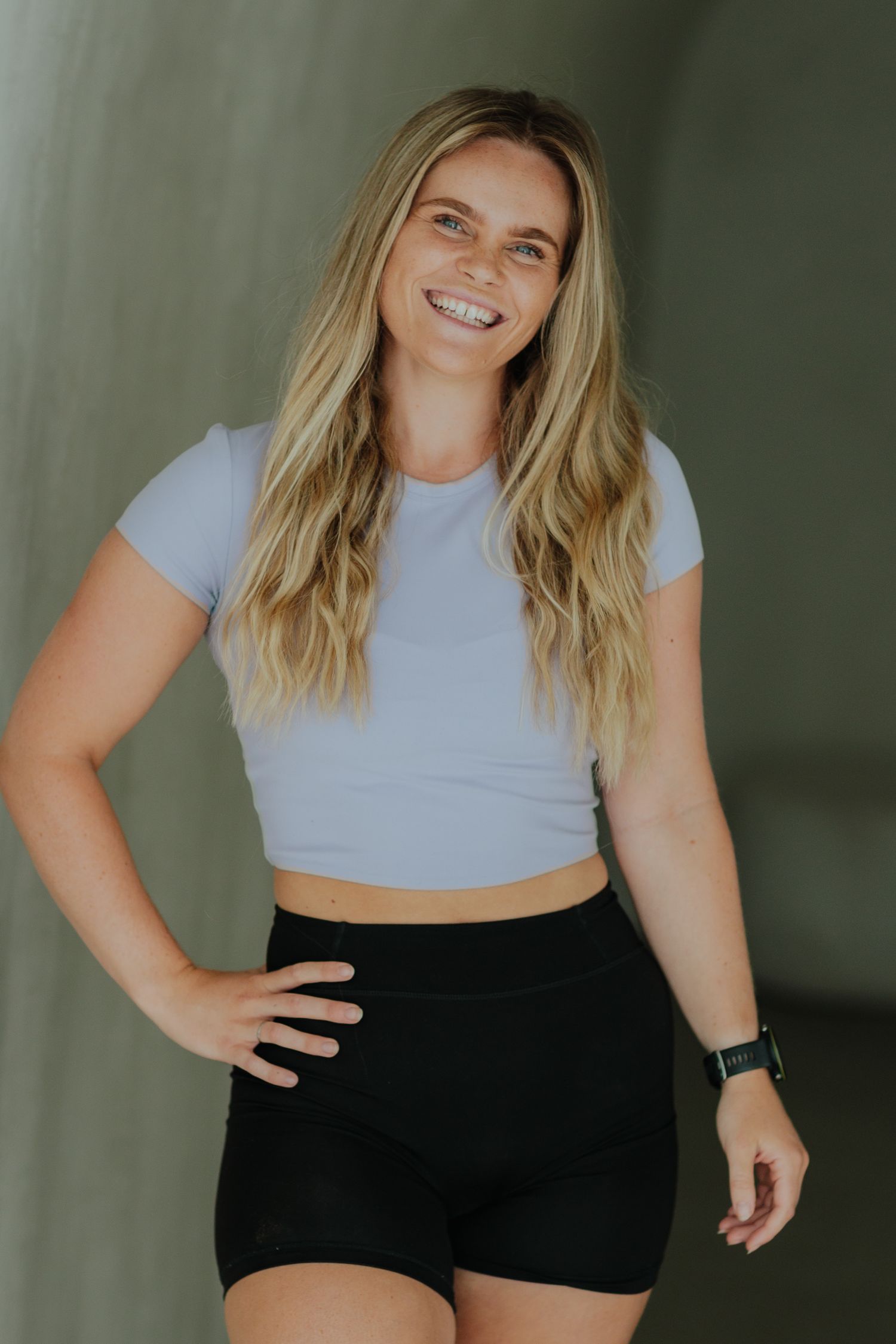 Smiling blonde woman in light blue crop top and black biker shorts posing confidently with hand on hip.