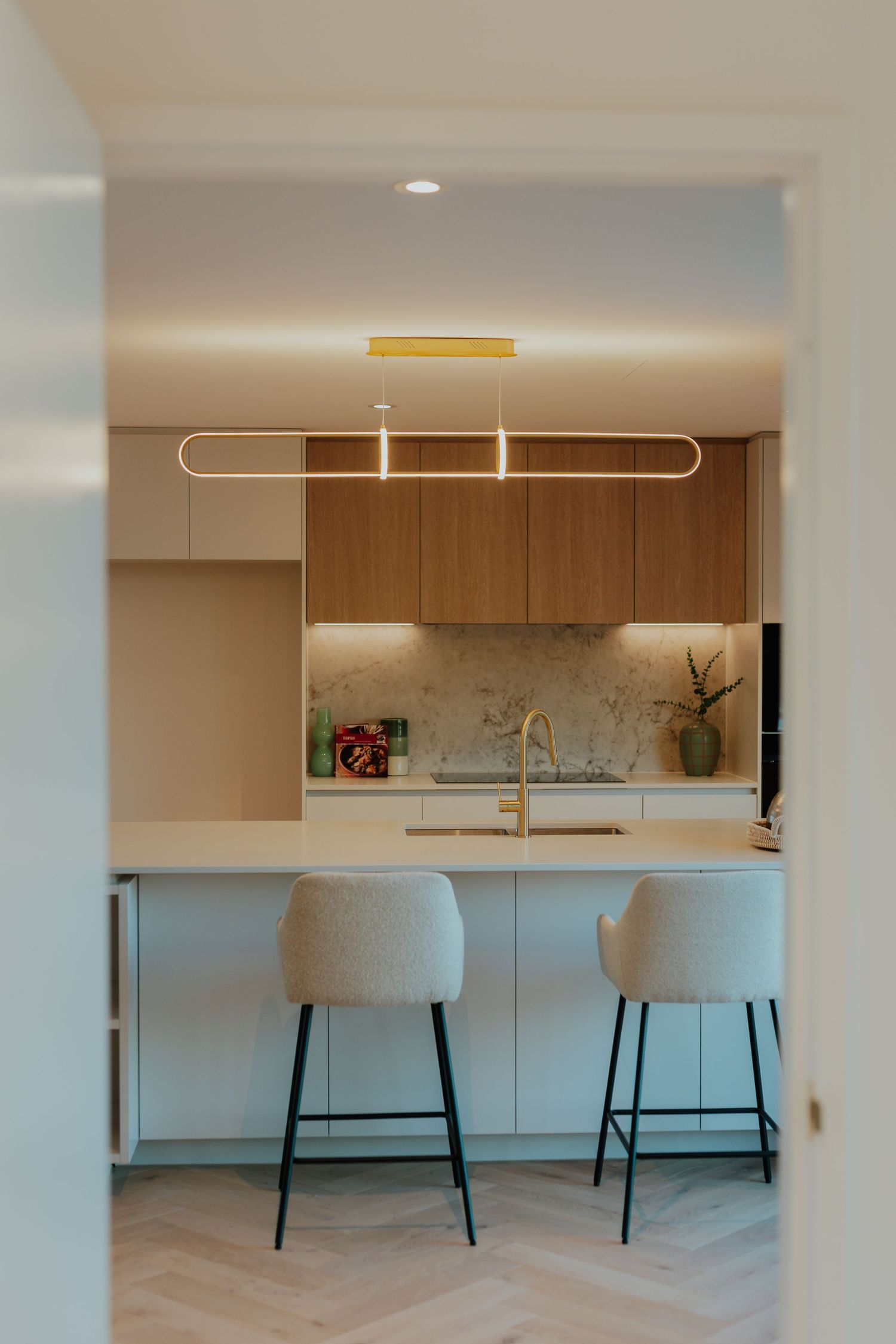 Elegant kitchen viewed through doorway with timber cabinetry, marble splashback, gold tap, and curved bar stools.