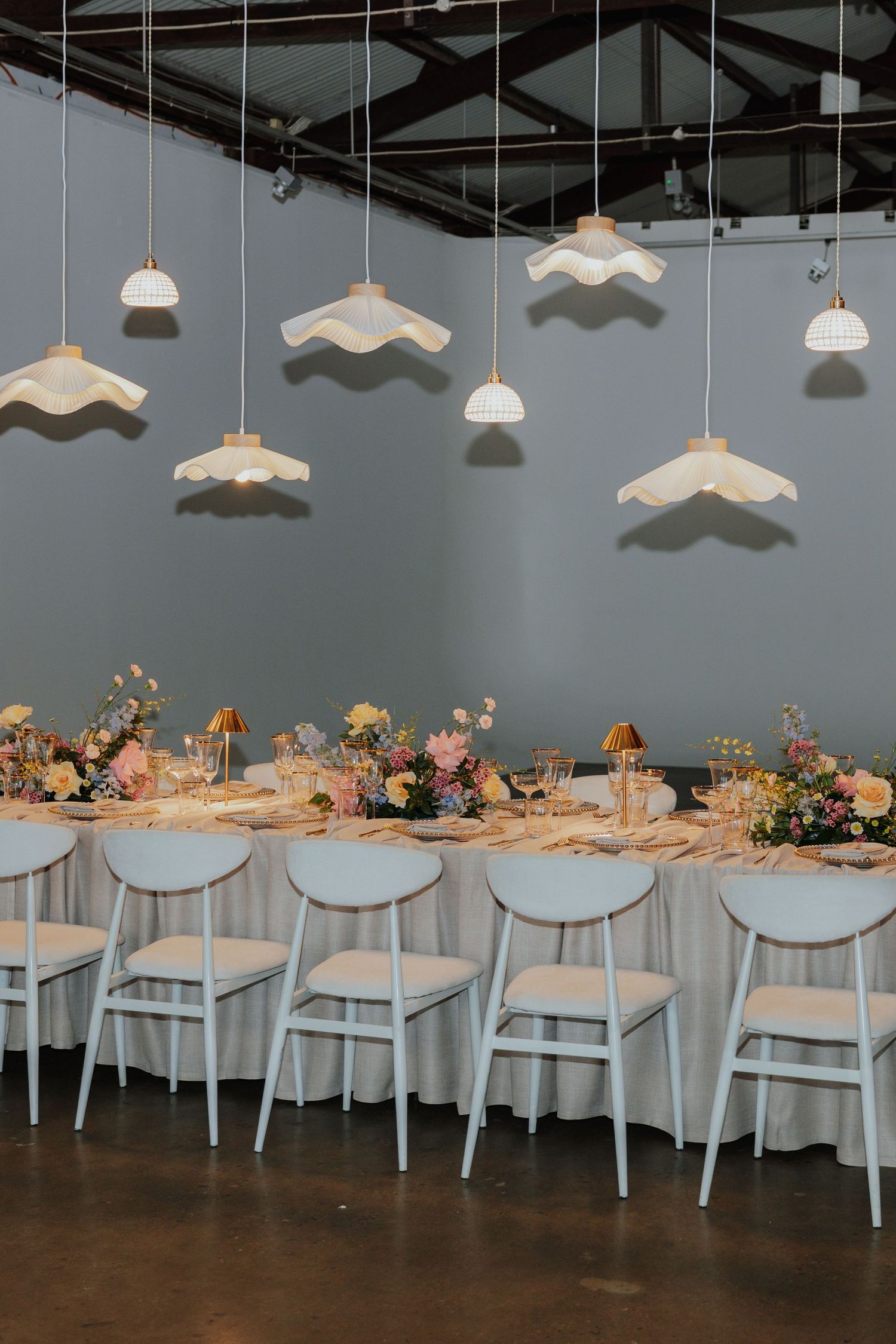 Long wedding reception table with light blue chairs, colorful floral arrangements, and wavy white pendant lights overhead.