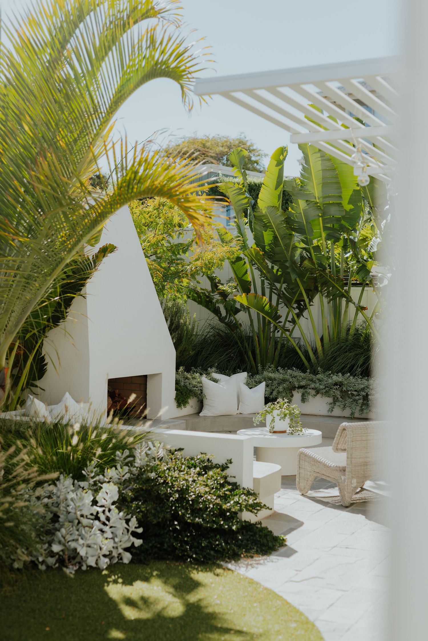 Lush tropical garden patio with white fireplace, curved bench seating, and white flowering shrubs framed by palm trees.