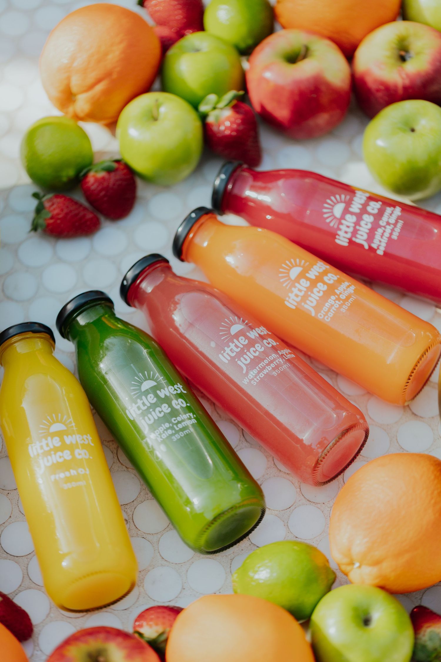 Colorful fresh fruit juices from Little West Juice Co. surrounded by apples and strawberries.