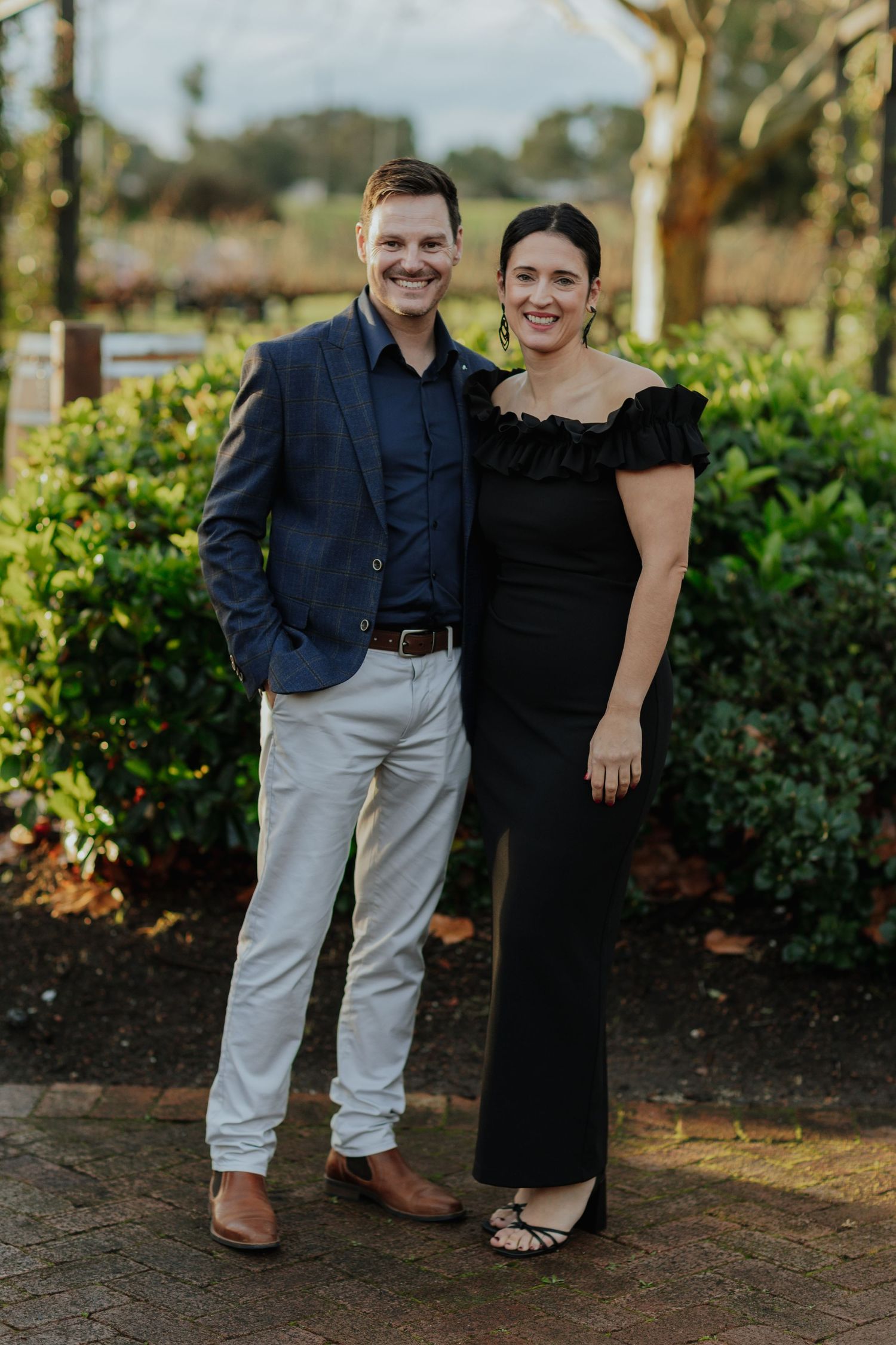 Smiling couple posing outdoors in formal attire, man in navy blazer and woman in black off-shoulder gown.