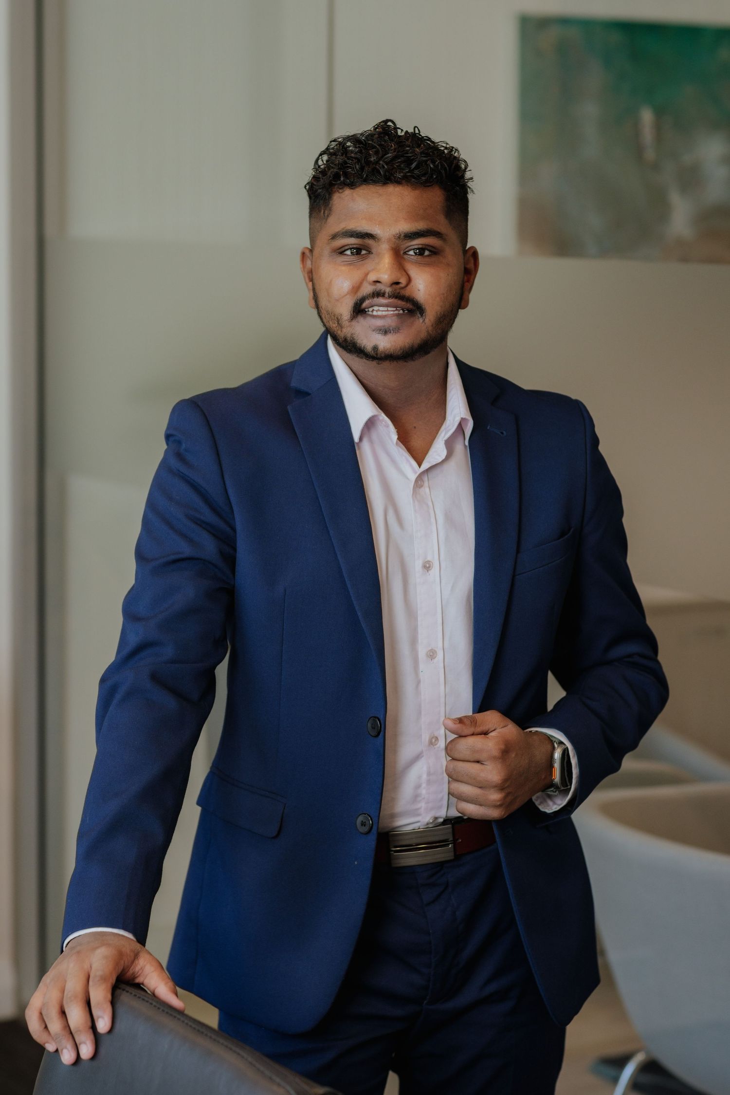 Confident young businessman in navy blue suit posing in a modern office environment.
