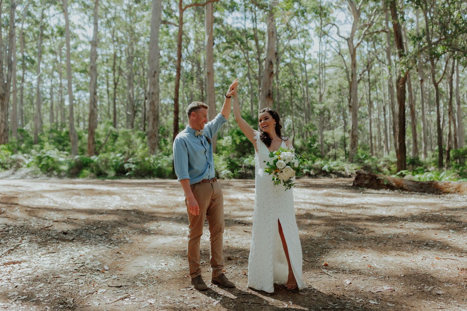 Bride and groom celebrate in sunlit forest clearing as groom raises bride's hand triumphantly.