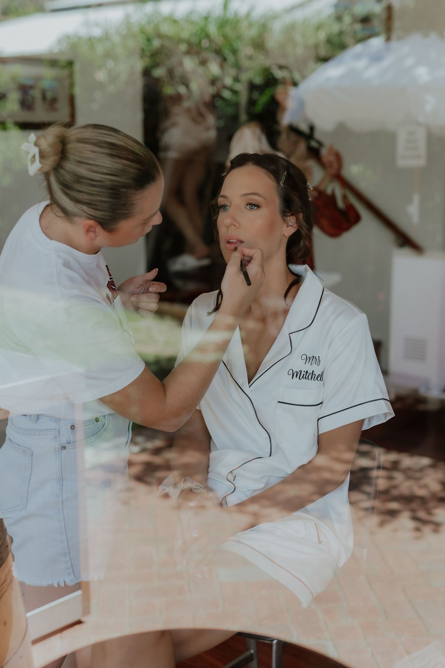 Makeup artist applies lipstick to brunette bride in white monogrammed pajamas during wedding prep.