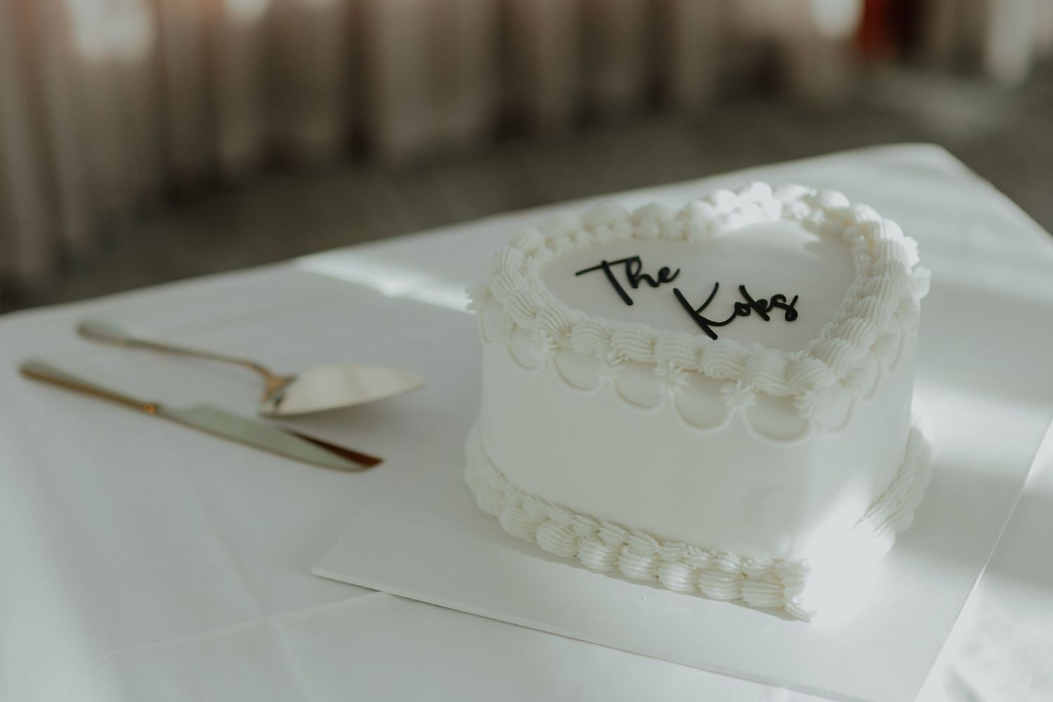 Elegant heart-shaped white wedding cake inscribed with The Kims on a white table.
