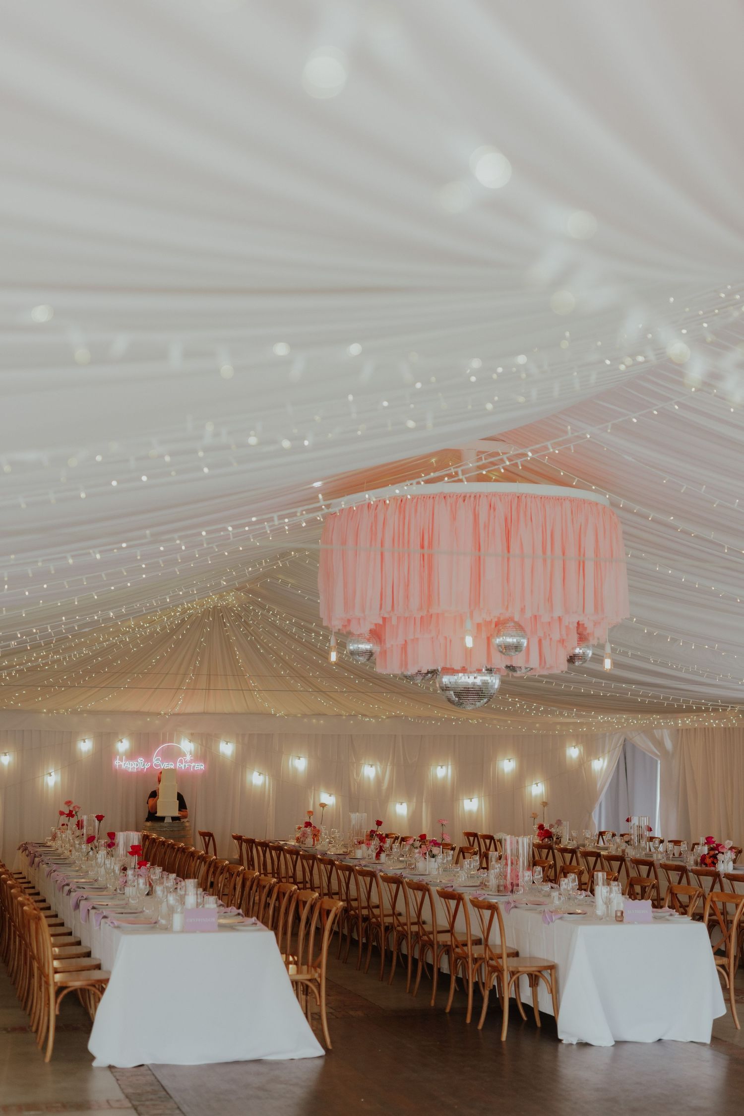 Elegant wedding reception marquee with fairy lights and pink chandelier over long tables.