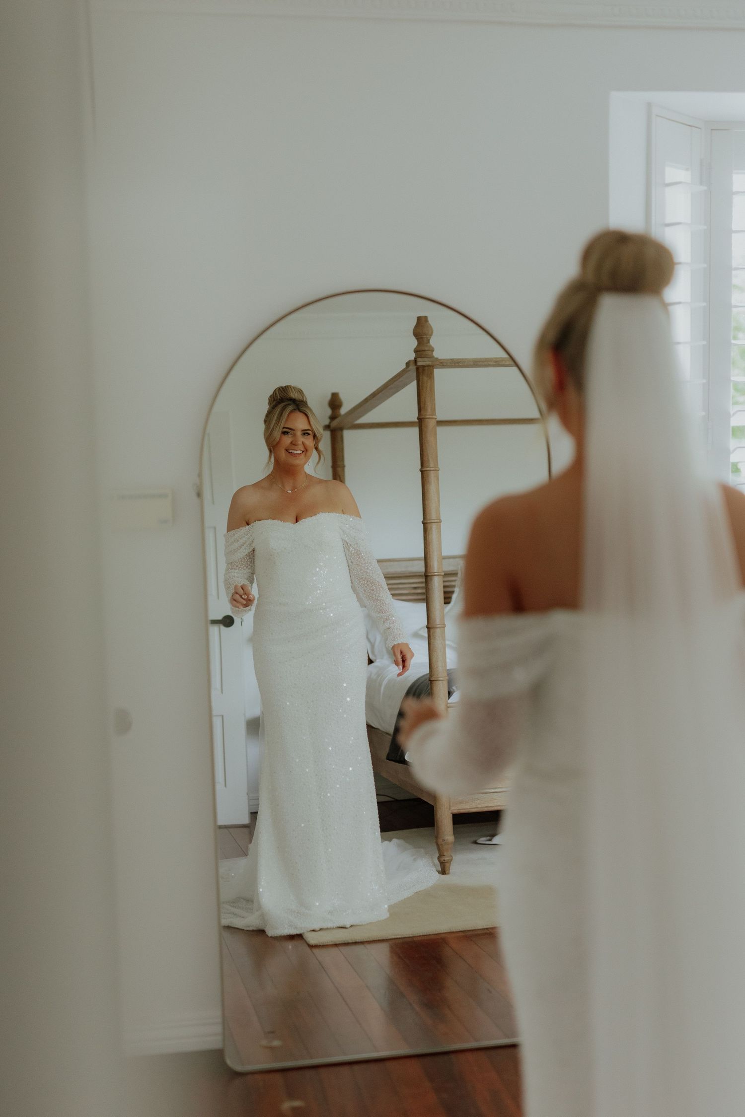 Elegant bride in off-shoulder white gown admires her reflection in large arched mirror.
