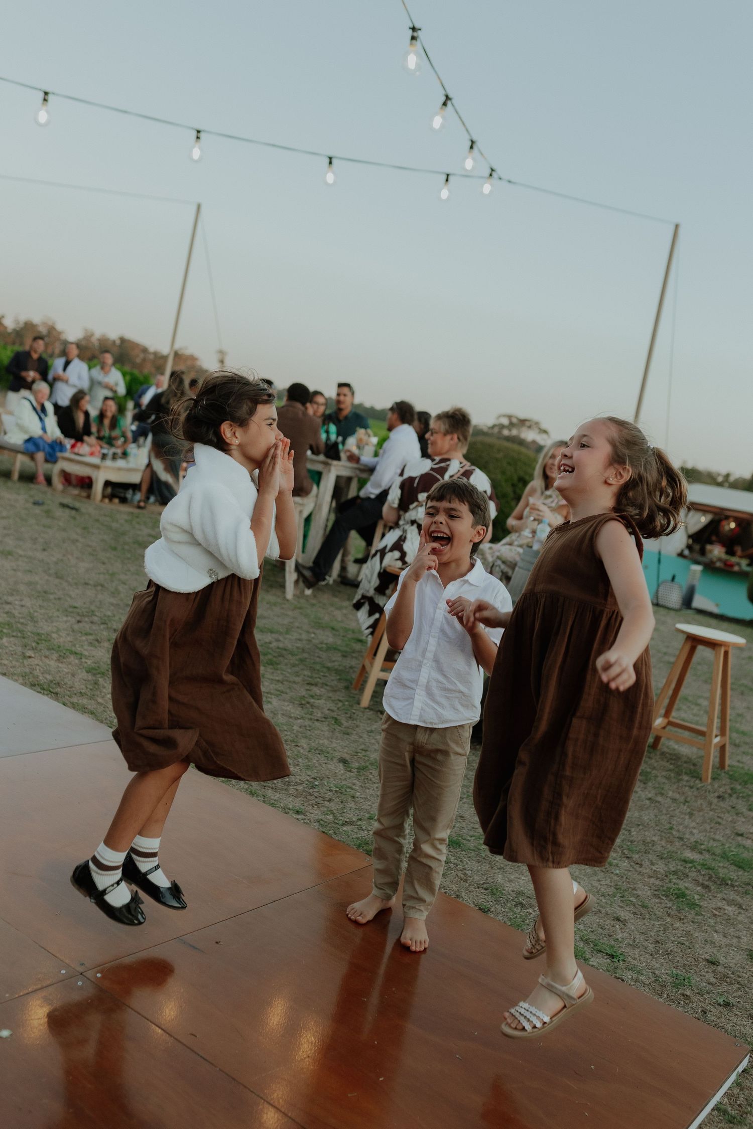 Three children laughing and dancing joyfully on outdoor dance floor at a twilight wedding reception.